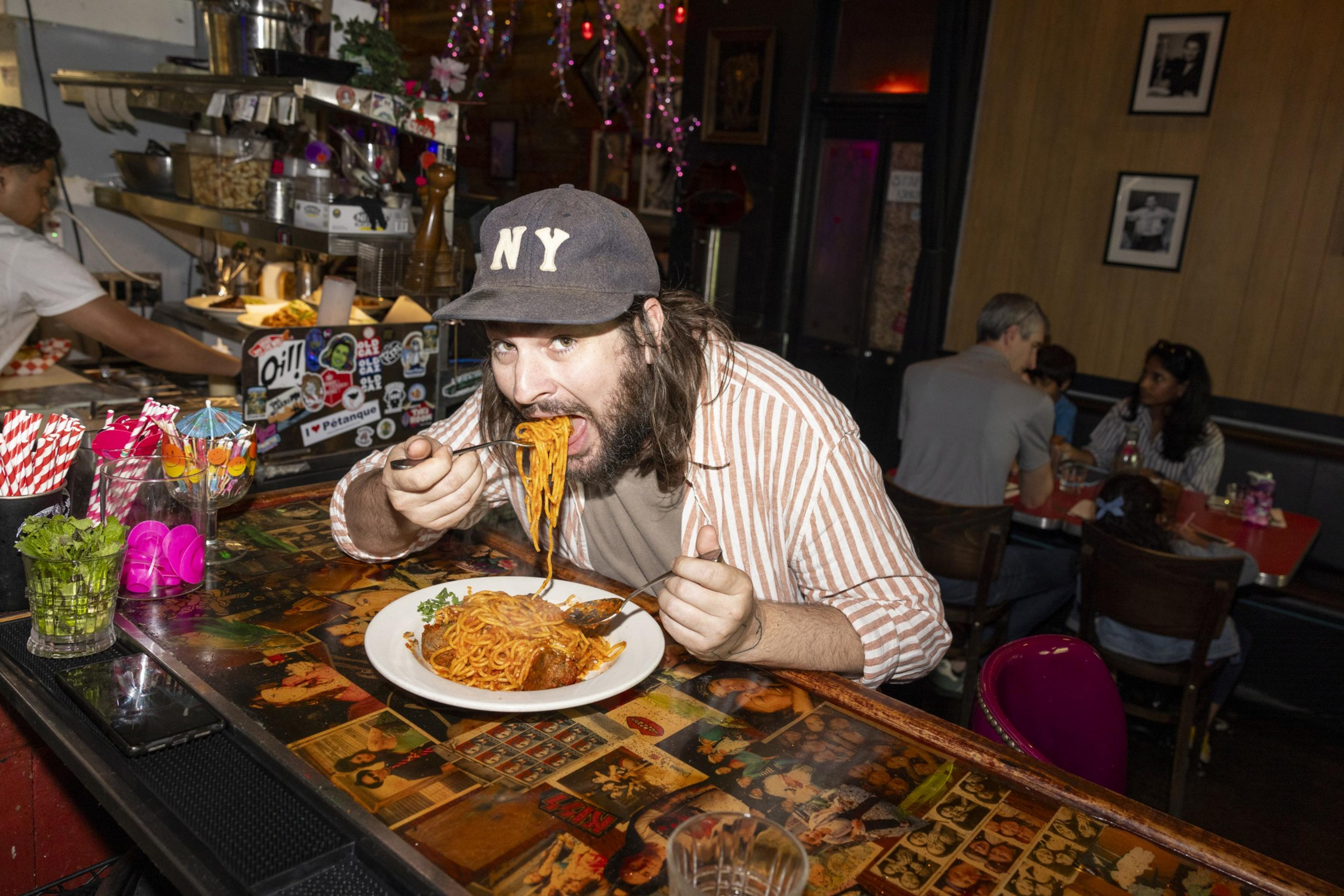 A man wearing a cap labeled &quot;NY&quot; is enthusiastically eating spaghetti at a colorful restaurant counter, with people seated in the background and a busy kitchen seen behind him.