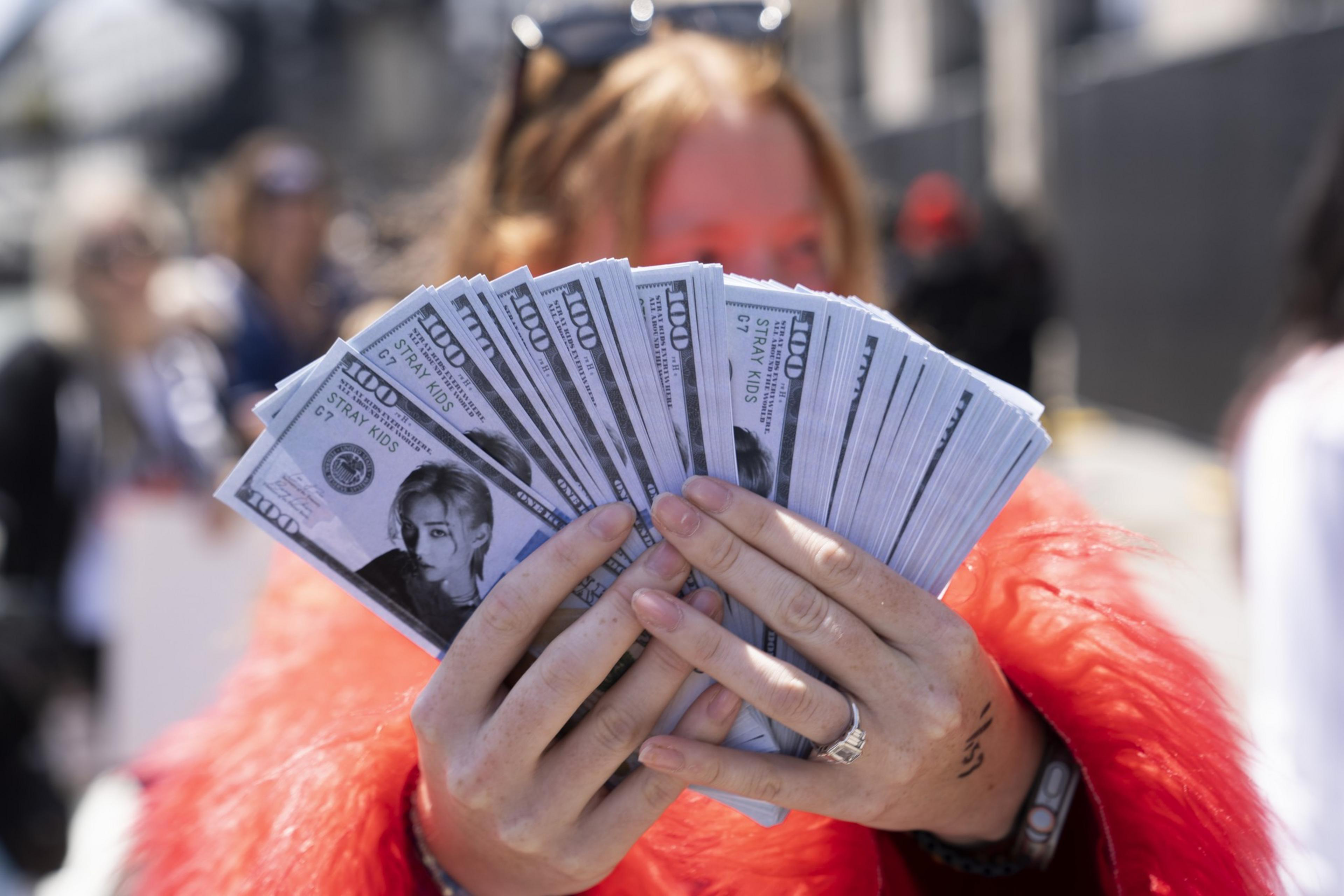 A person in a red jacket holds multiple fans of novelty bills featuring a portrait labeled &quot;Stray Kids.&quot; The background is blurred with indistinct figures.