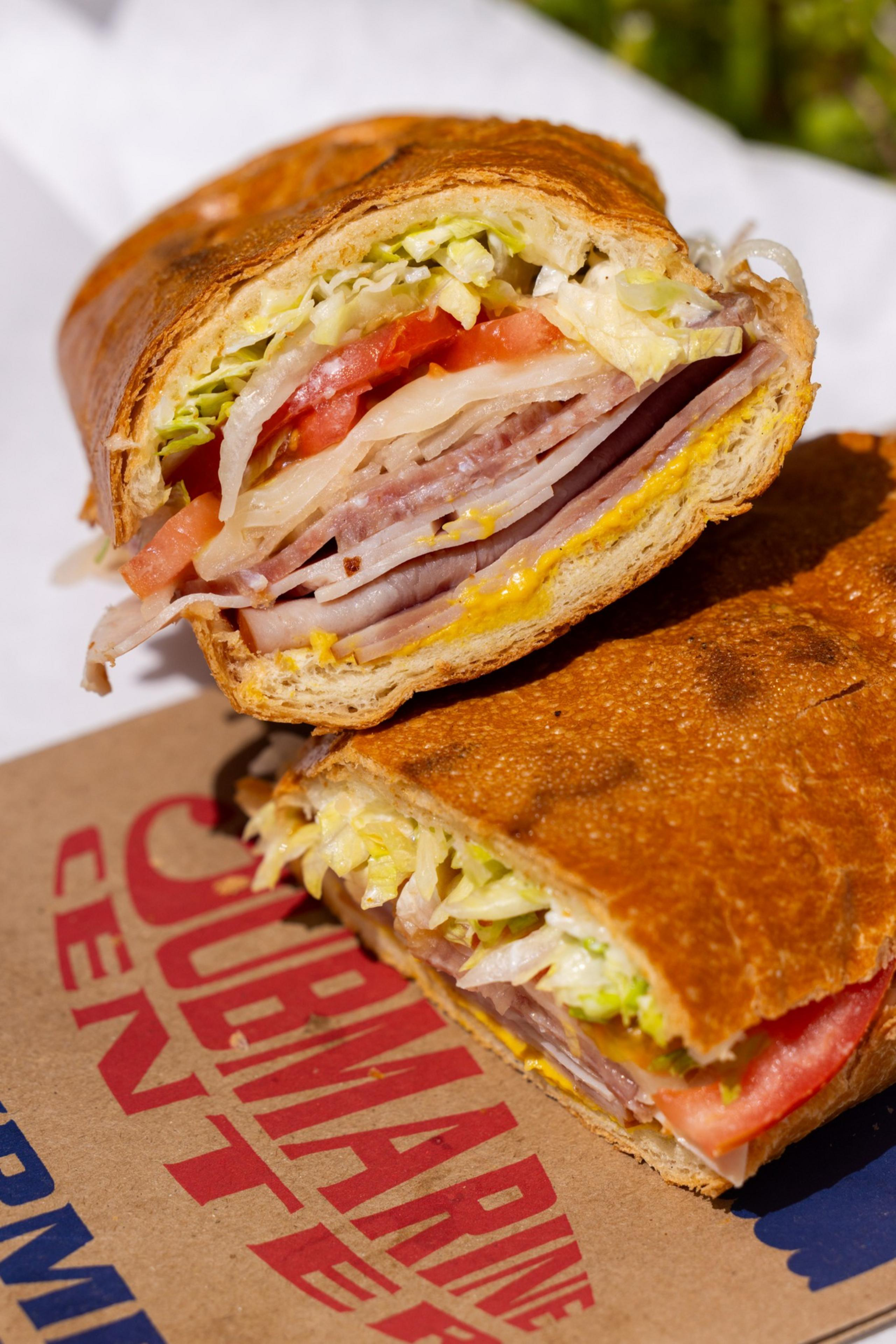 A sub sandwich with layers of lettuce, tomato, meat, and cheese is neatly cut in half, placed on a cardboard surface with printed red and blue text.