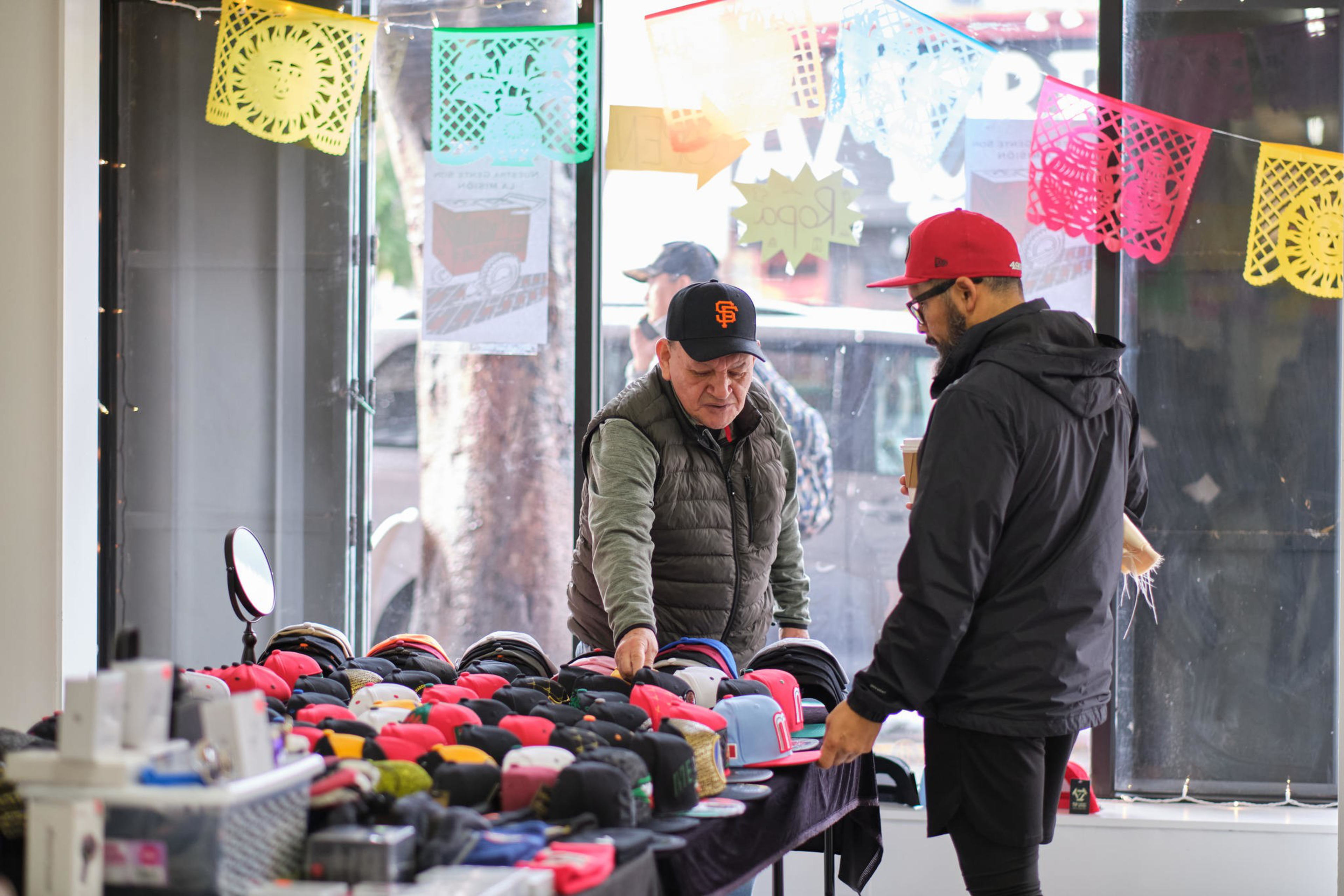 Two men stand by a table covered with various hats inside a room with colorful decorations.