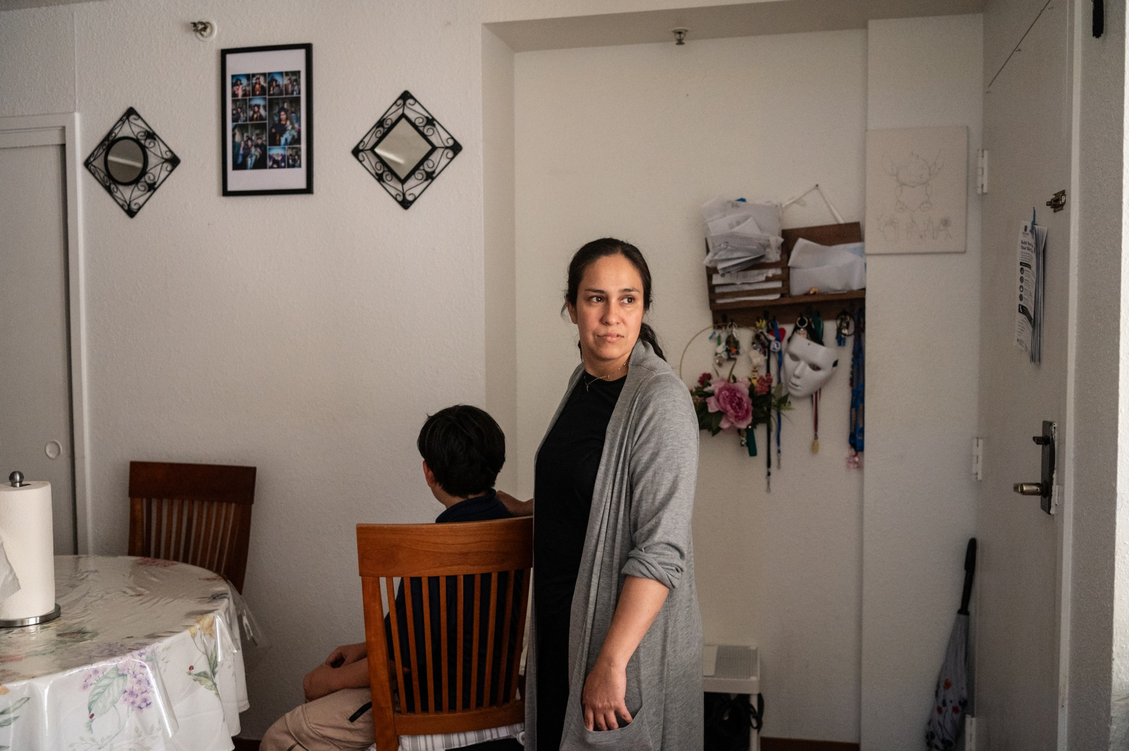 A woman in a gray cardigan looks to the side while a seated boy faces away in a modest room with wall decor, a table, and hanging items.