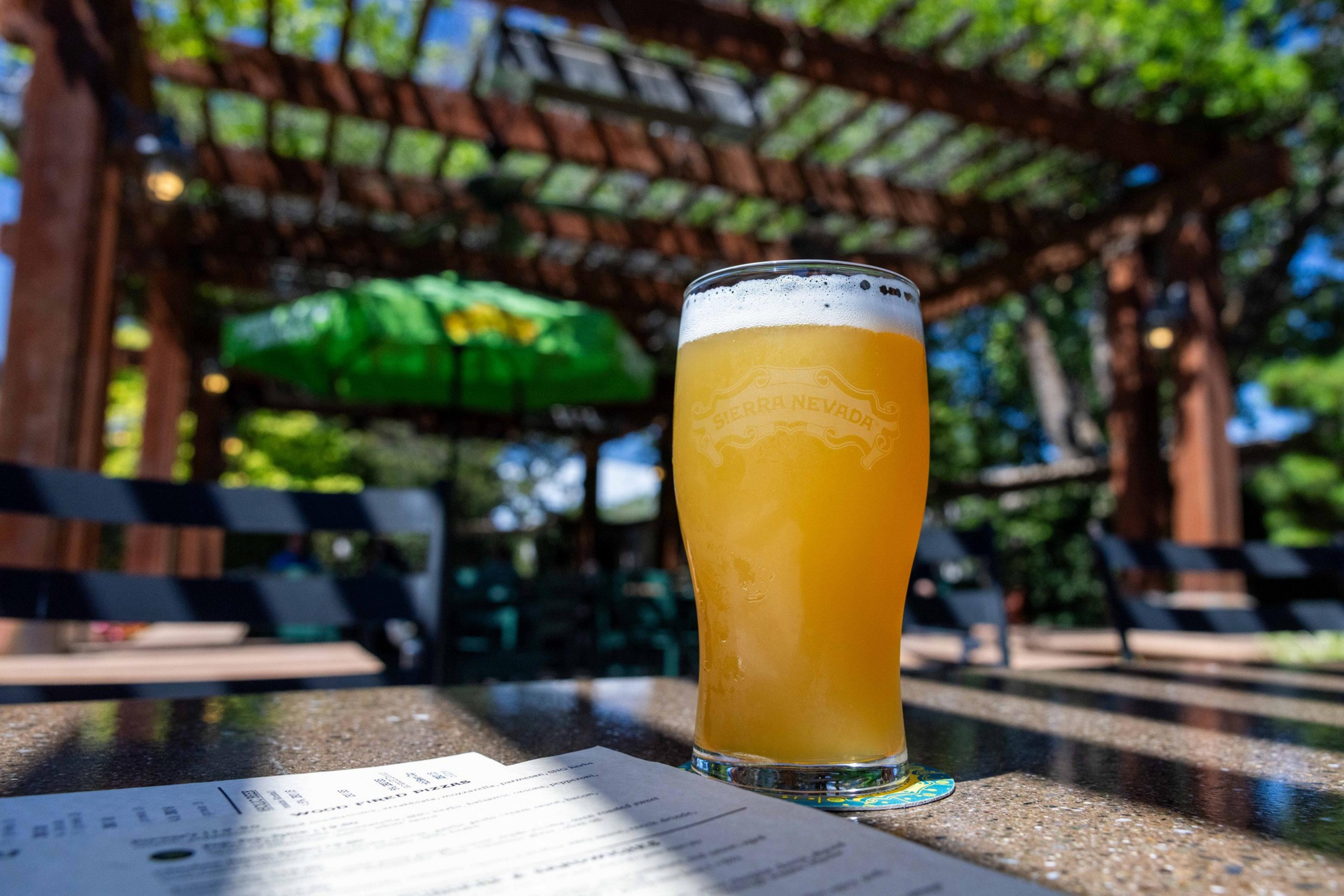 A frothy, orange-tinted beer in a Sierra Nevada glass sits on a sunlit table, with a blurred outdoor setting and menu in the background.