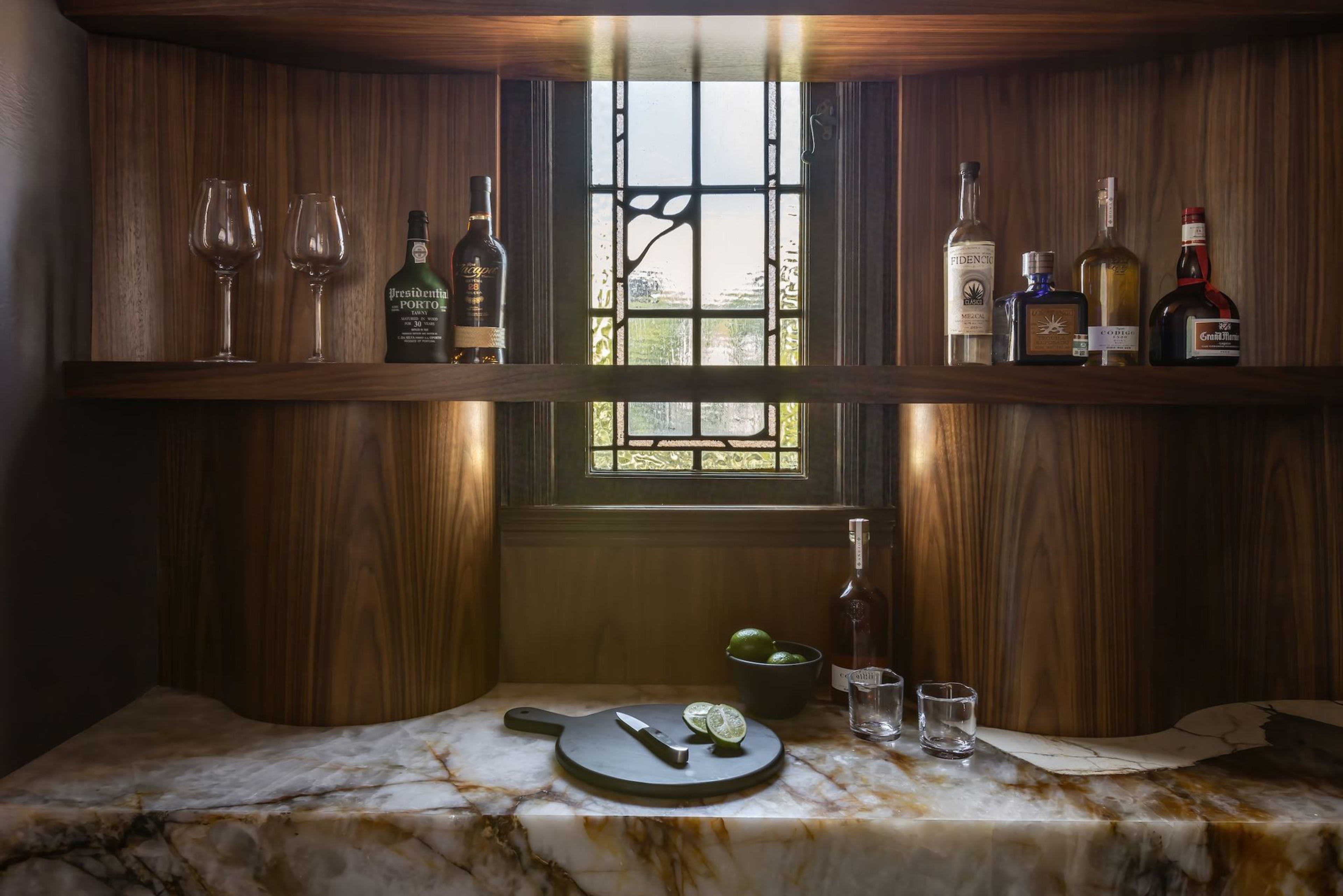 Wooden bar shelves hold wine glasses and assorted liquor bottles; a marble countertop below has a cutting board with sliced limes, a bowl, two glasses, and a bottle.