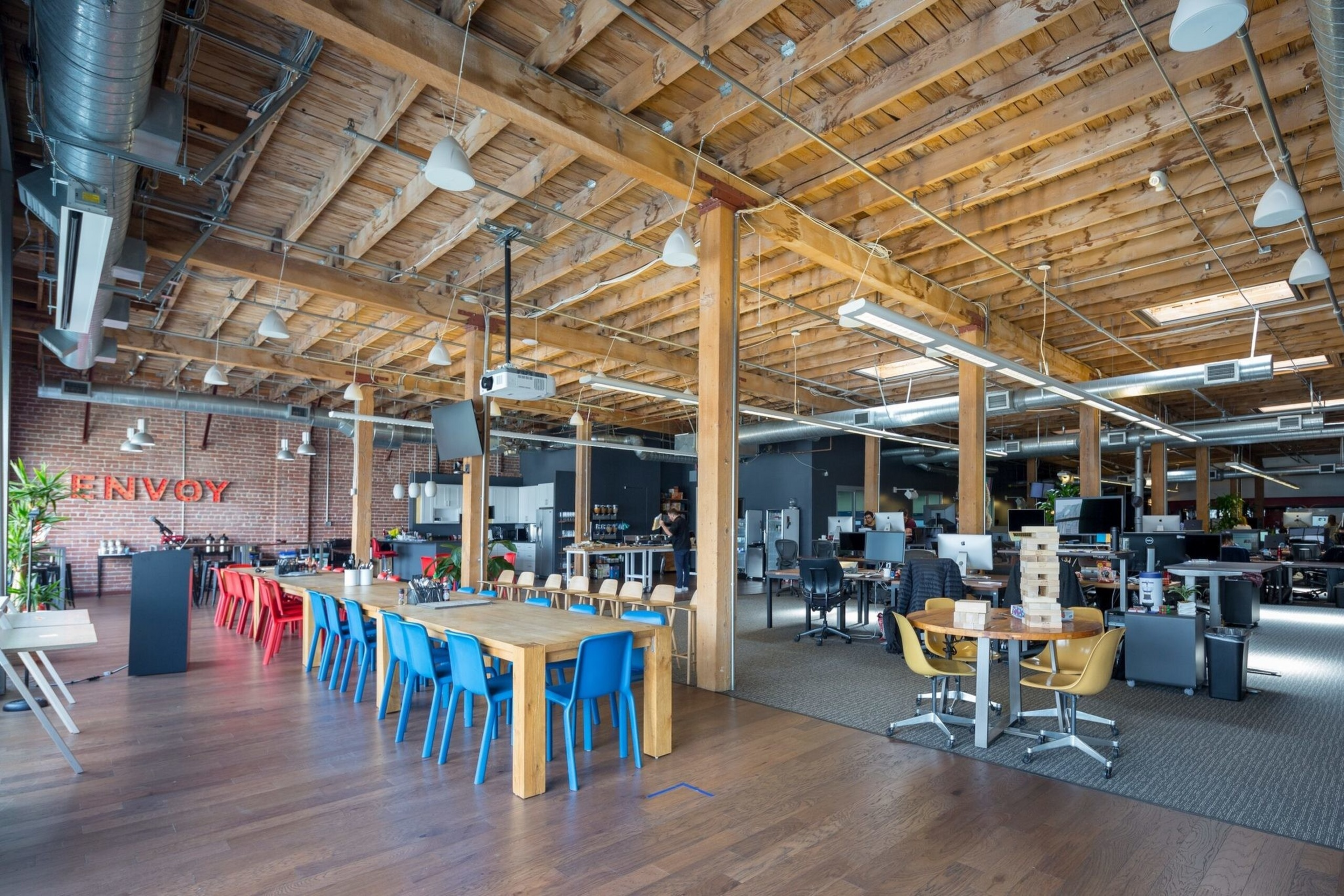 An open office space with wooden beams and floors, tables with colorful chairs, computer workstations, a kitchen area, and a brick wall with "ENVOY" in red letters.