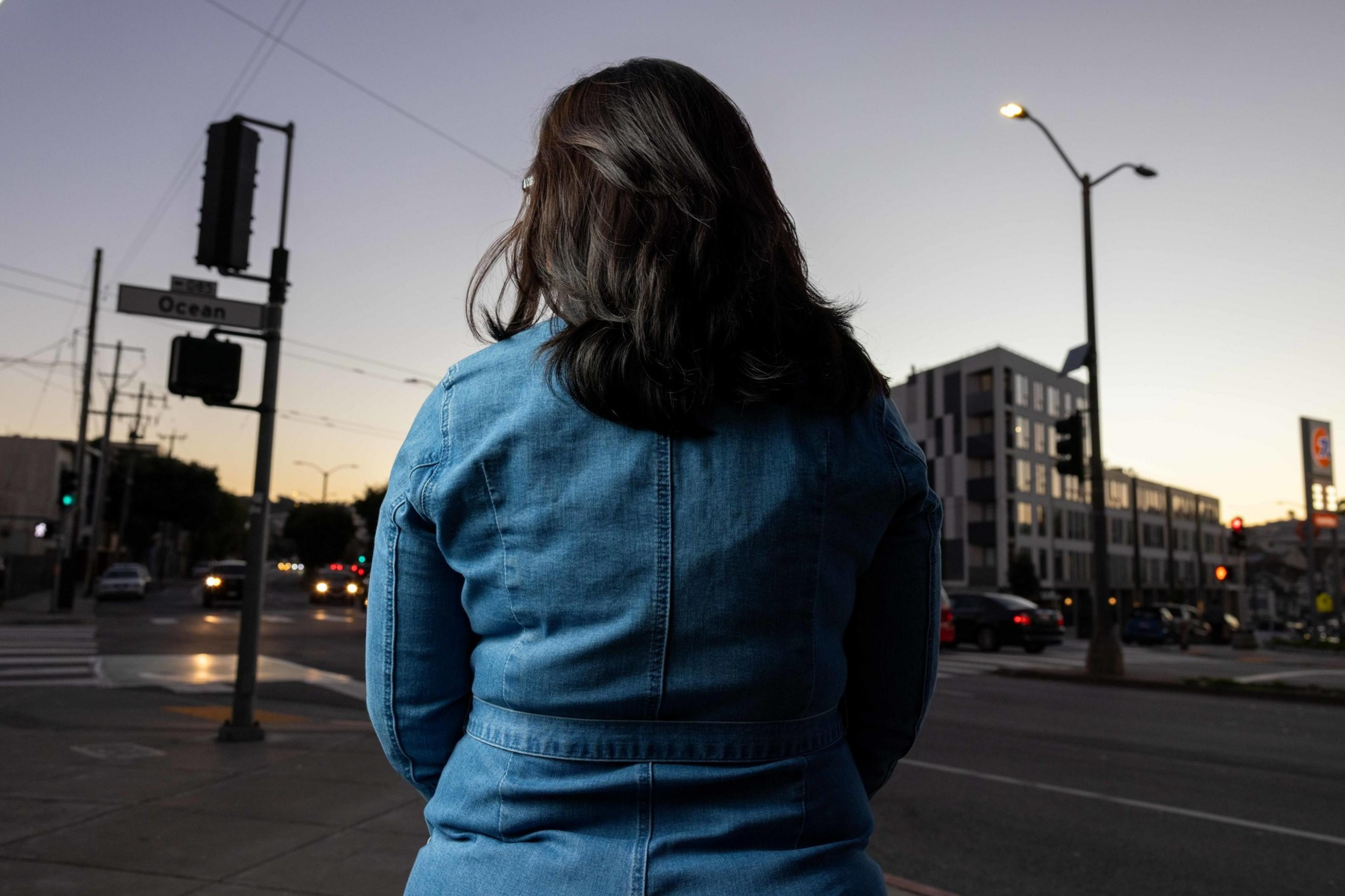 A woman with long dark hair, wearing a denim jacket, stands on a city street at dusk. Streetlights and cars are visible, with buildings in the background.
