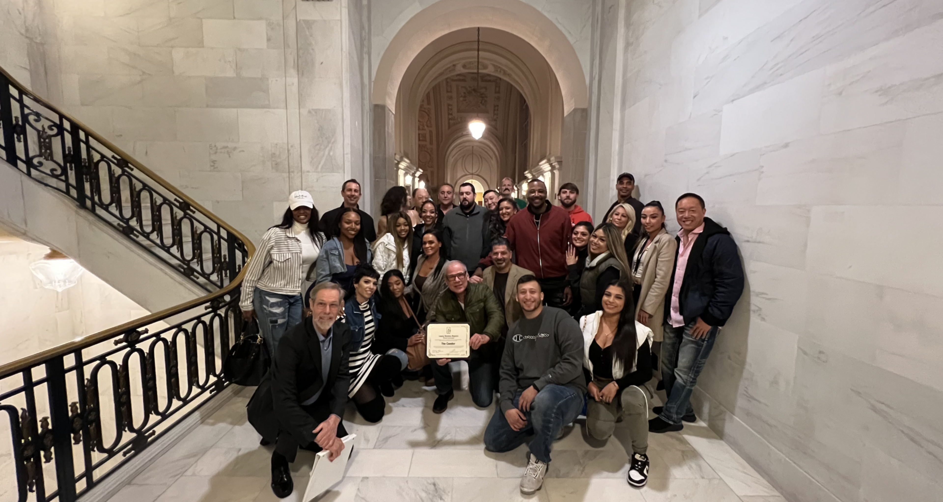 A diverse group of 28 people poses smiling in a marble hallway, with one person holding a certificate in the center.