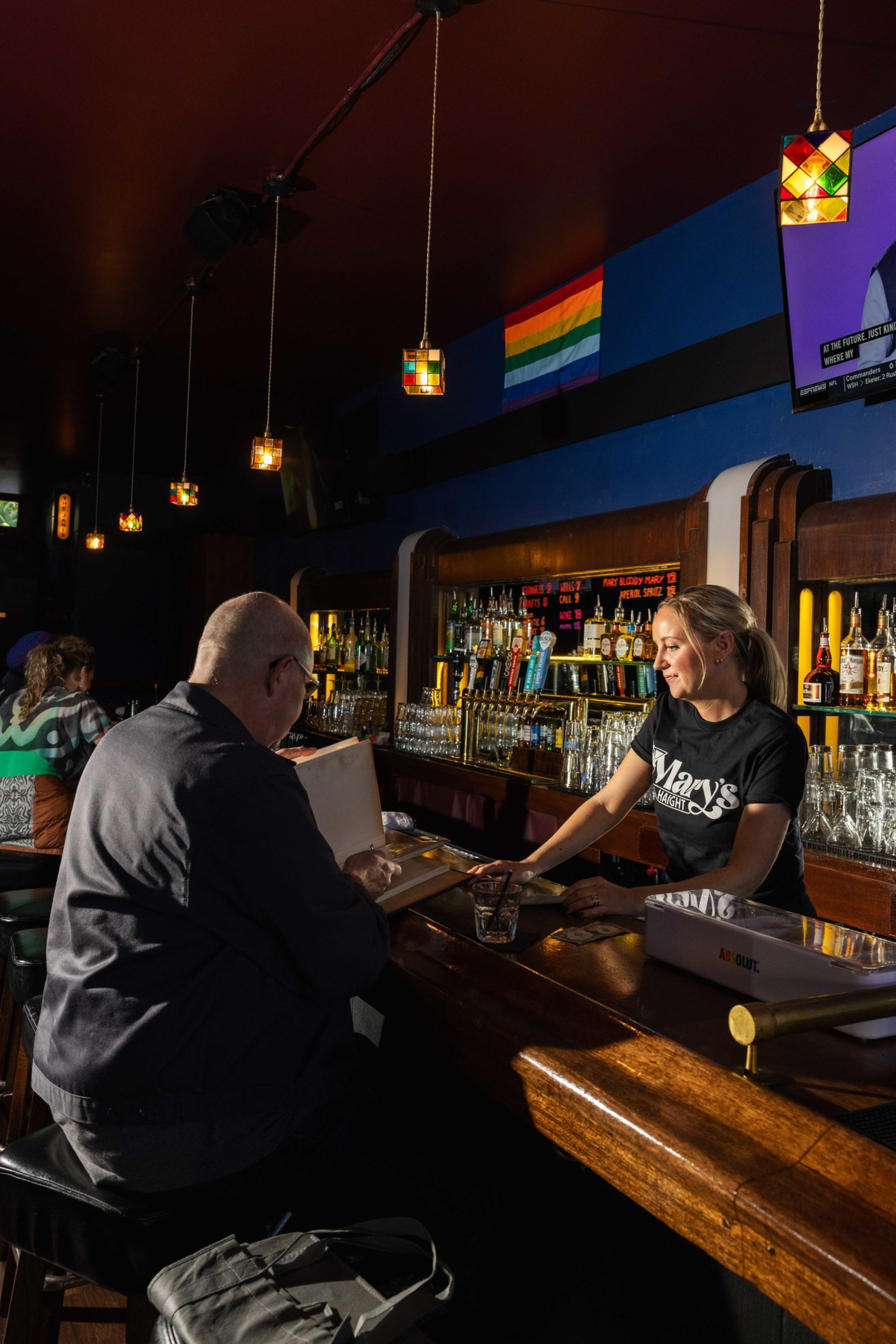 A bartender wearing a “Mary’s” shirt serves a seated customer at a dimly lit bar with hanging lanterns and a rainbow pride flag on the wall.