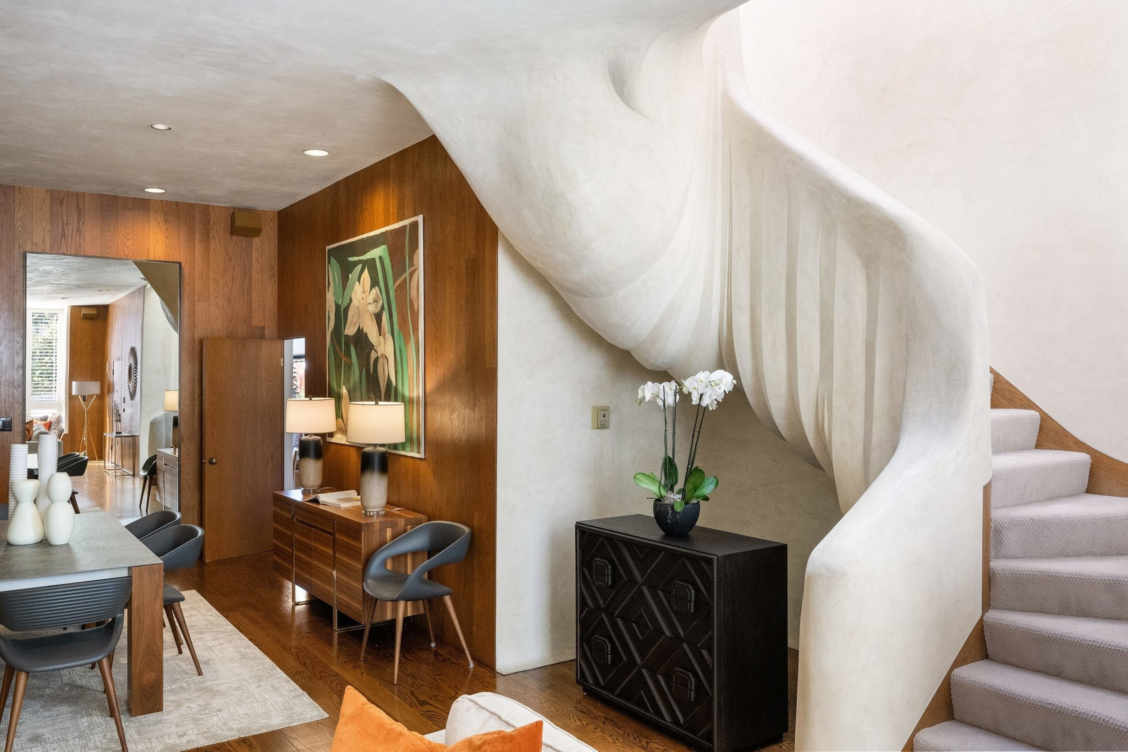 The image shows a modern interior with a sculptural curved staircase, wood-paneled walls, a dining area, and decor including a vase of white flowers on a dark cabinet.