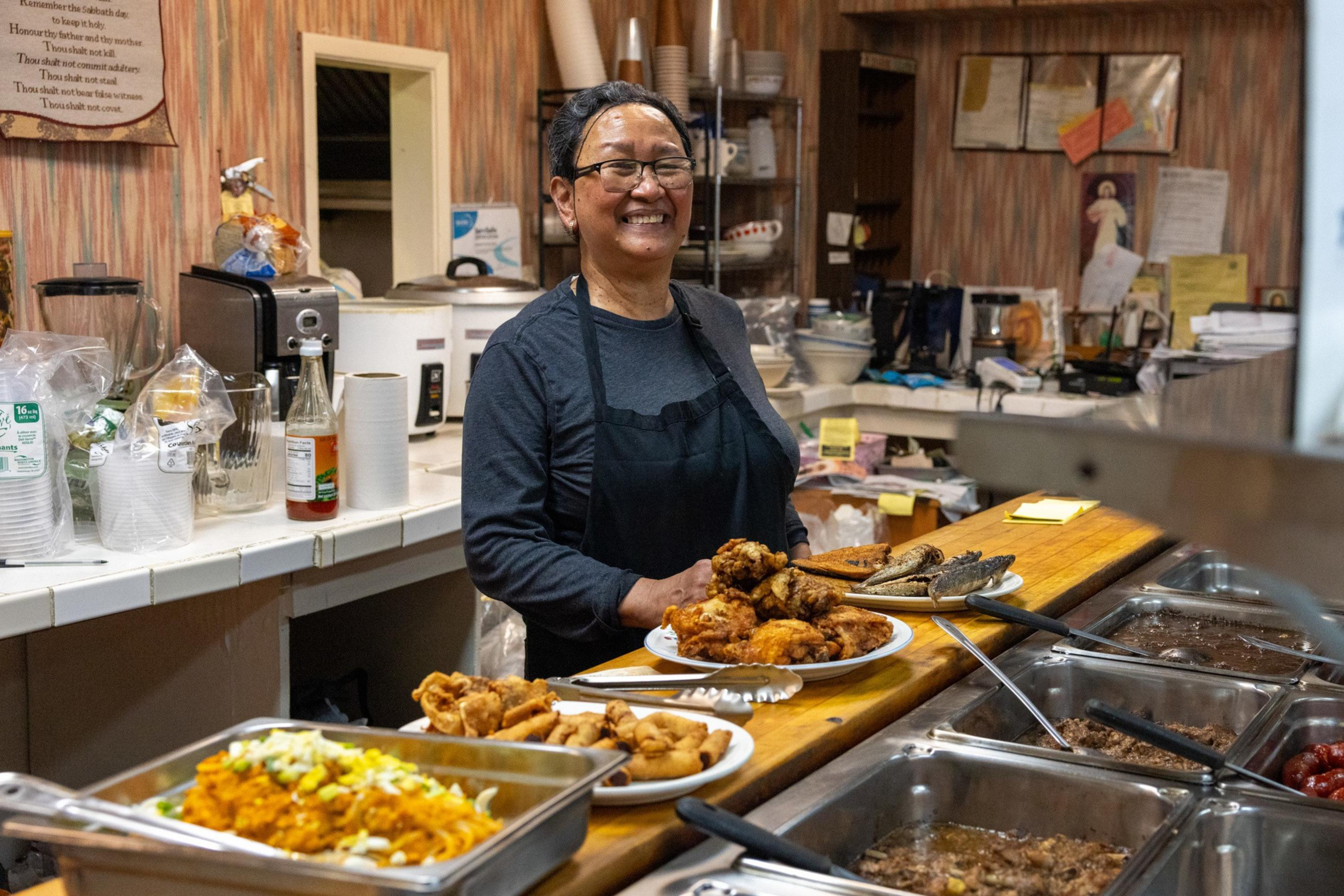 A smiling woman wearing glasses and an apron stands behind a counter with plates of fried chicken, fish, and trays of various hot dishes in a cozy kitchen.