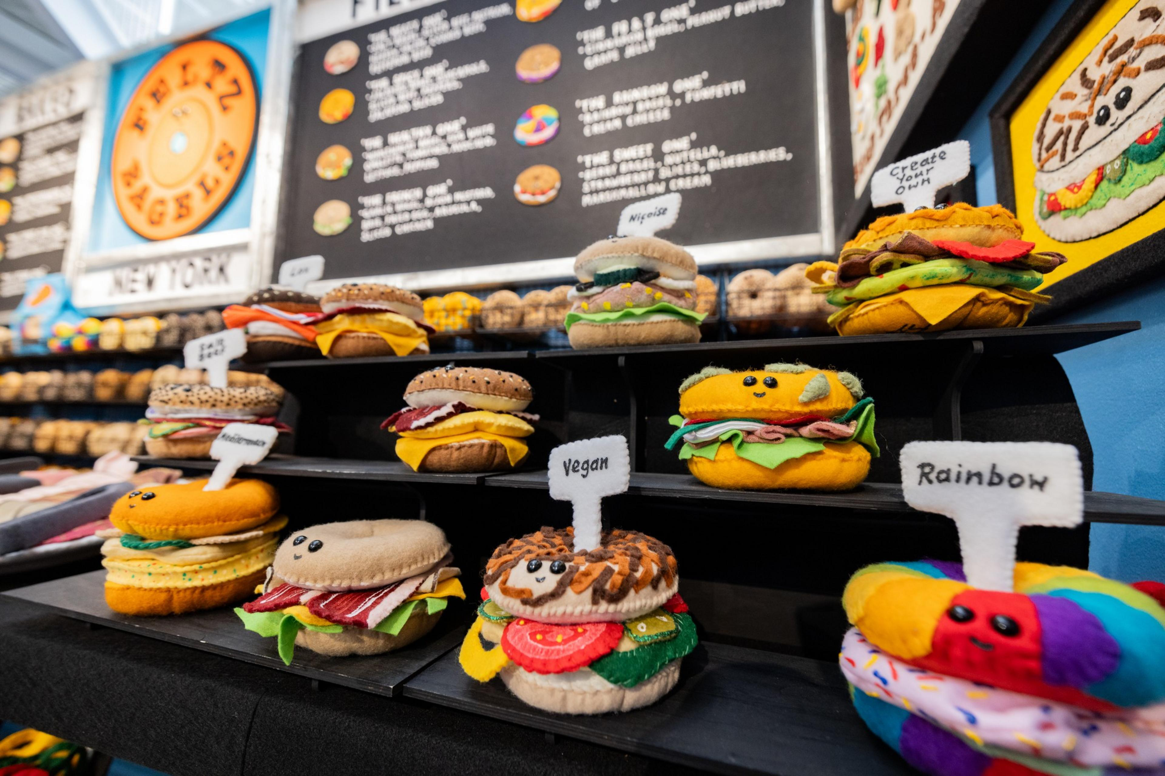 A row of bagels made out of felt stands on a shelf in front of a menu.