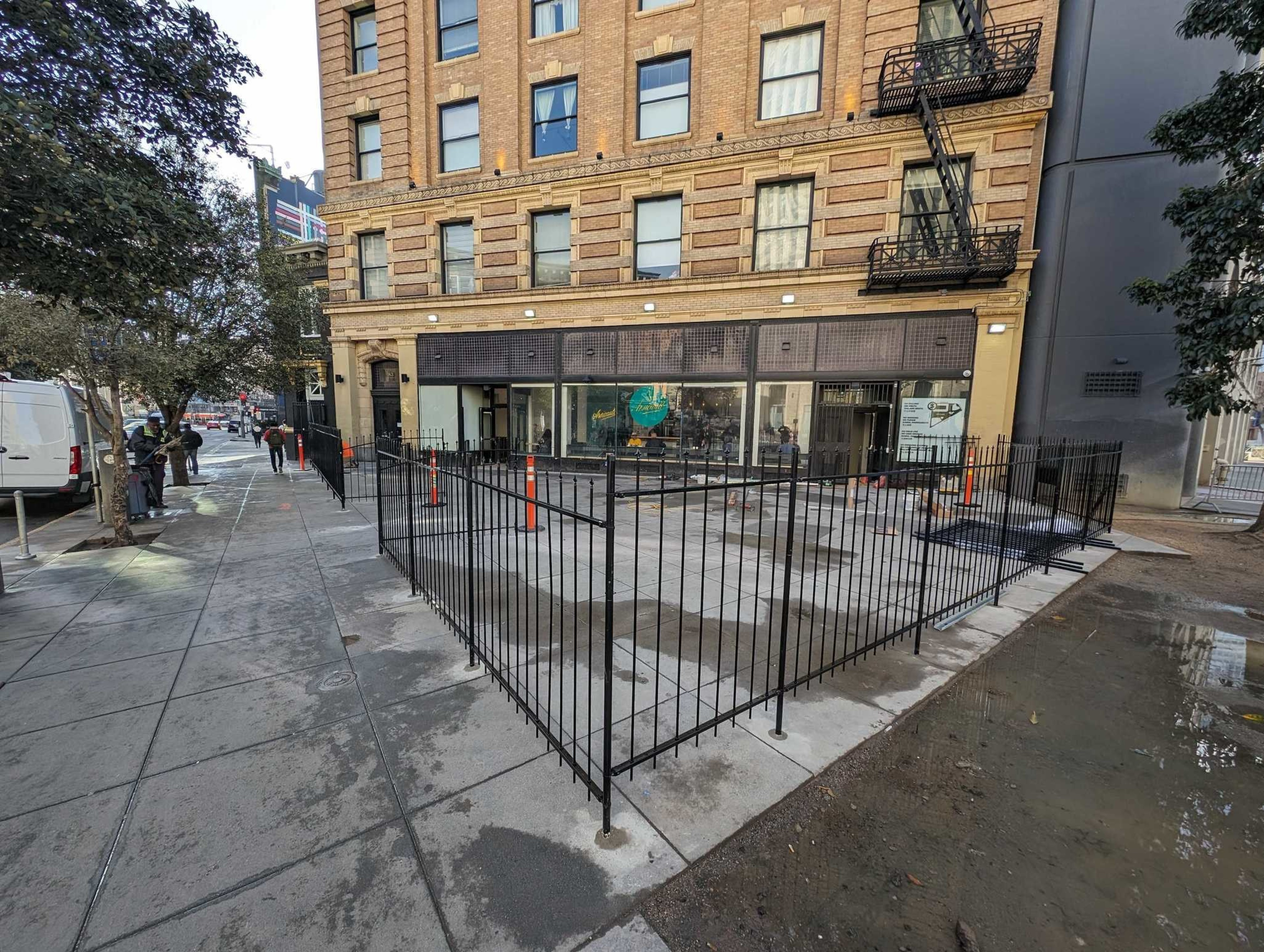 Urban street view with a traditional building on the left, metal fencing, trees, and walking pedestrians.