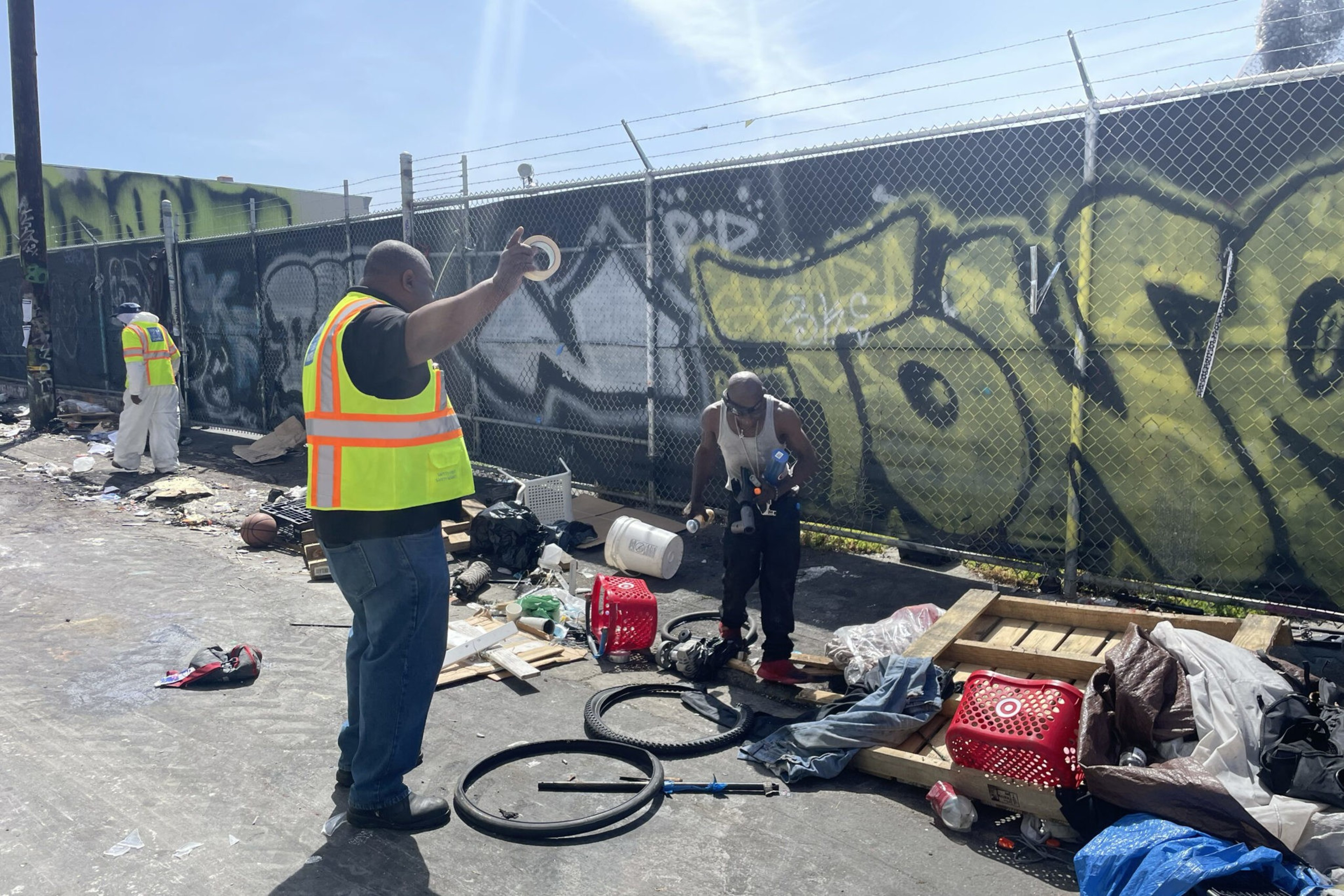 Two men, one in a yellow safety vest and another in a white tank top, are sorting through scattered trash and debris near a graffiti-covered fence.