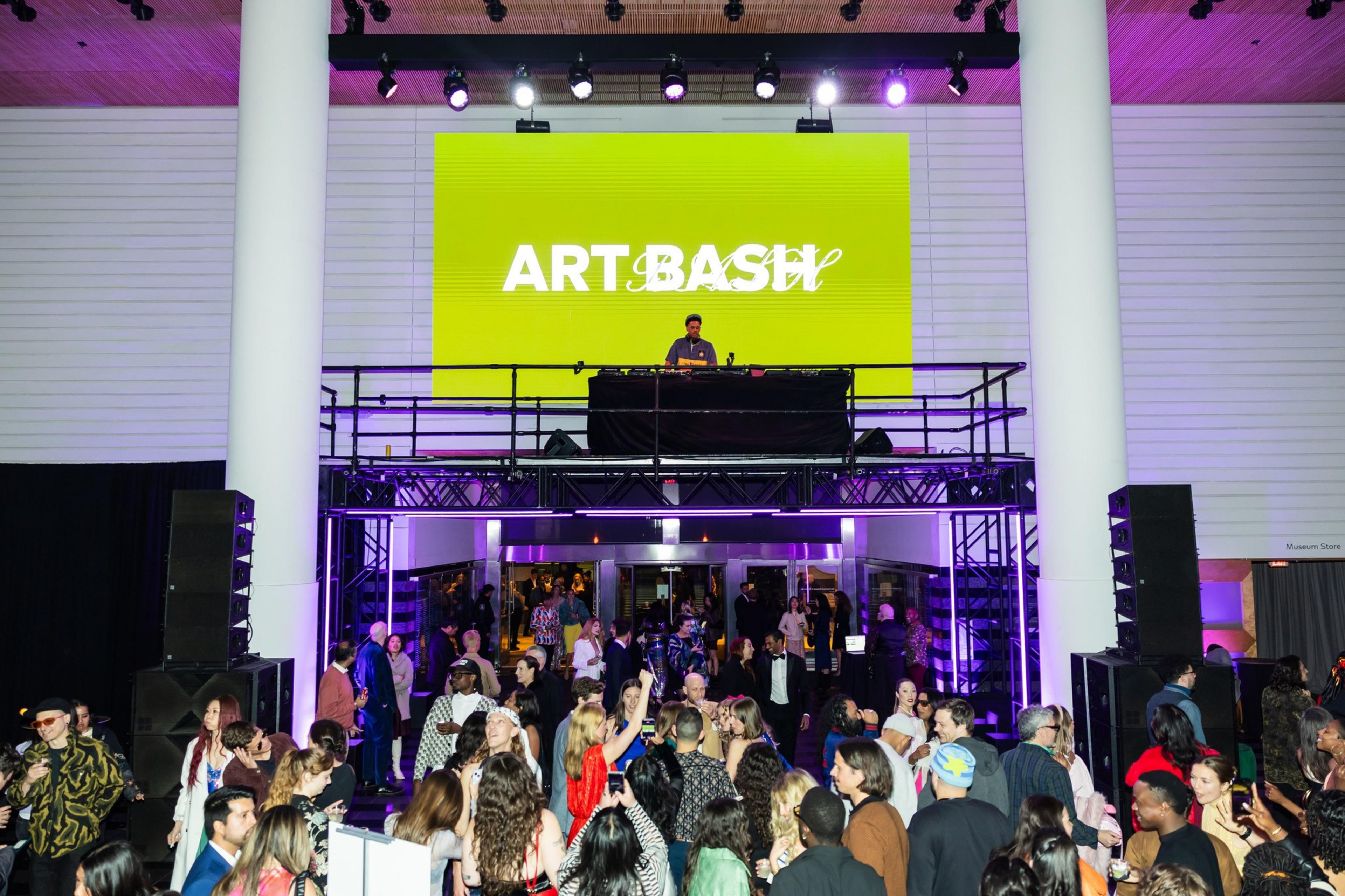A DJ performs on an elevated stage beneath a large bright green screen displaying “ART BASH” as a crowd of people mingles and dances below.