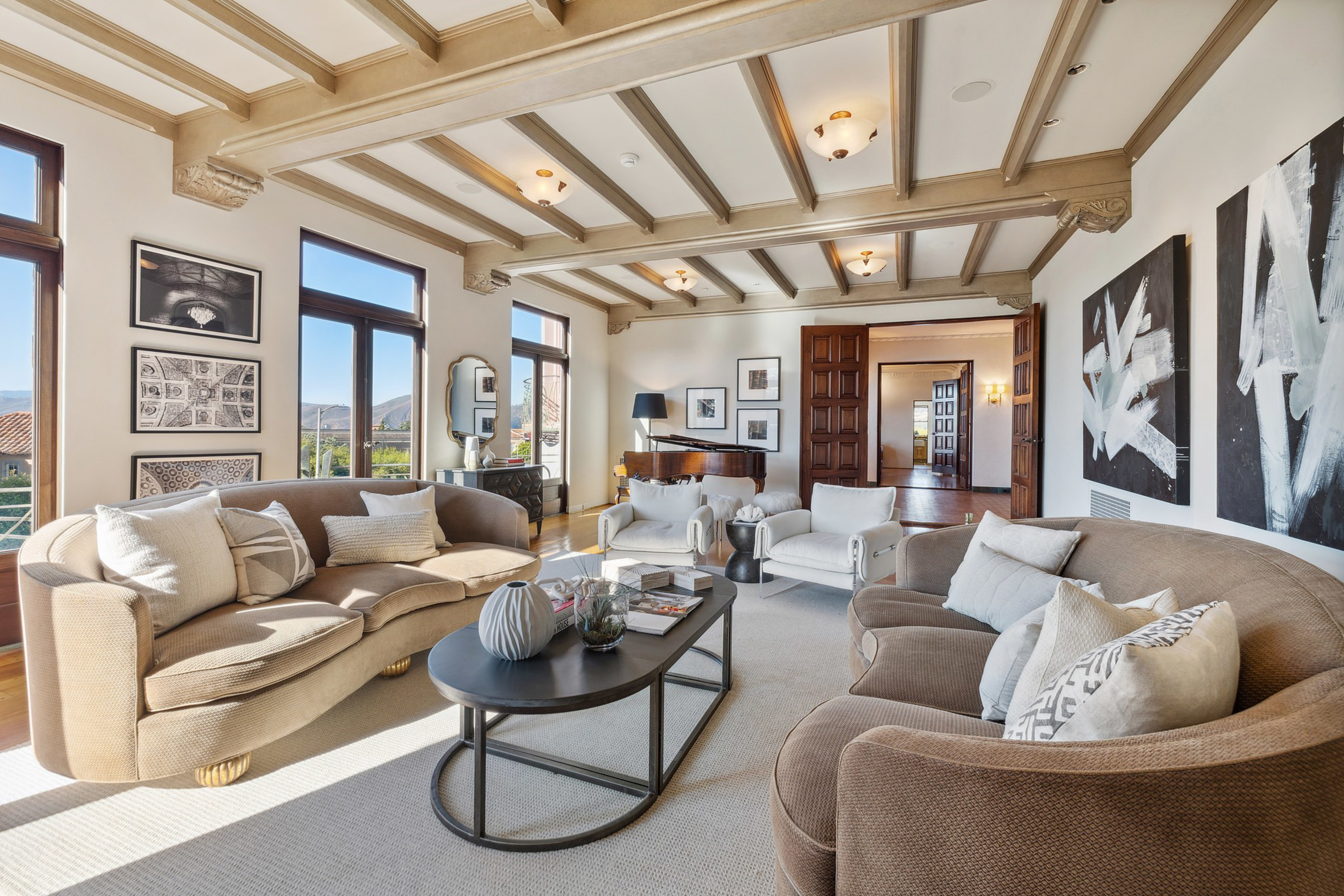 Two curved beige sofas and two white armchairs surround a black oval coffee table in a bright room with large windows, abstract art, and a grand piano.