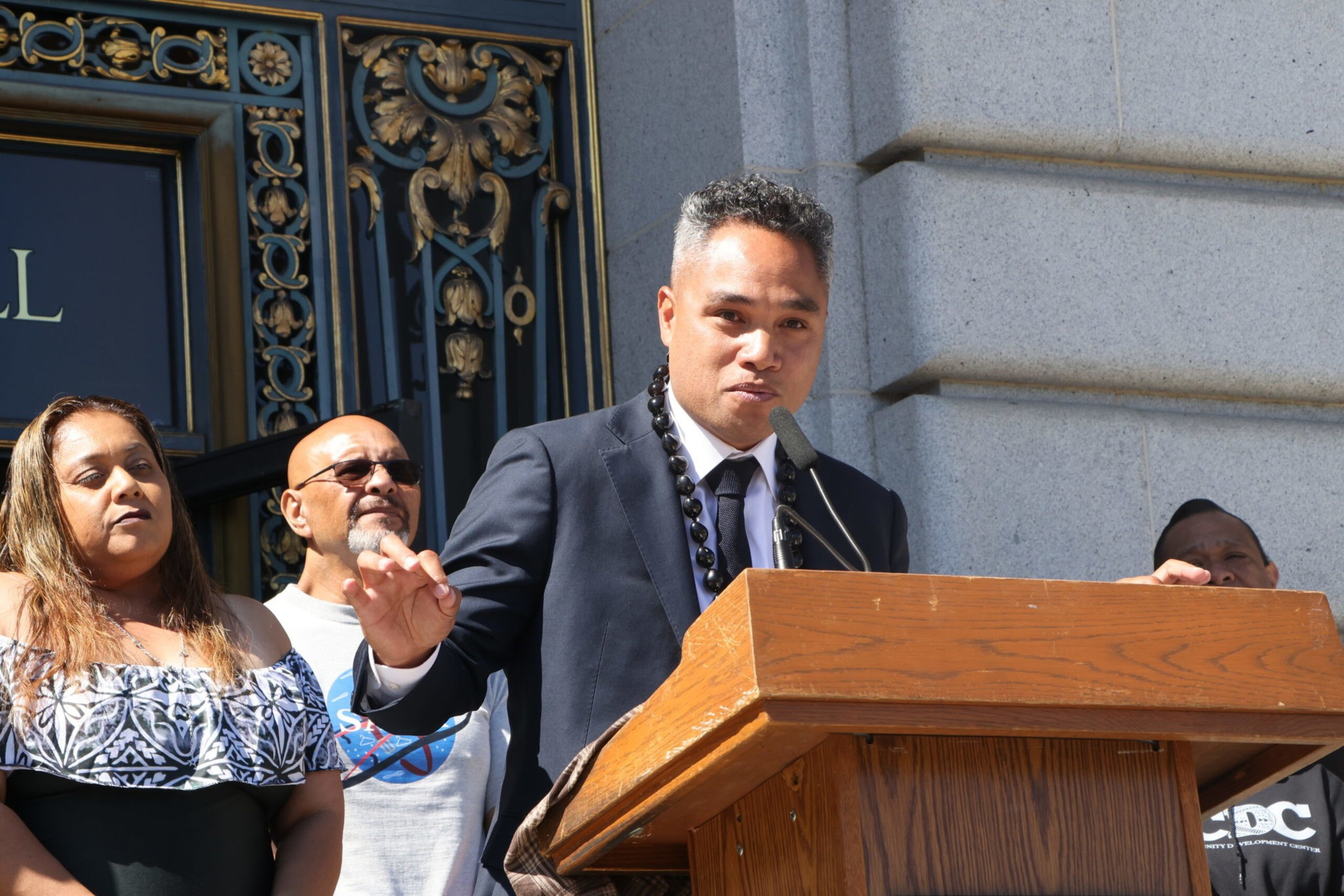 A man in a suit and lei speaks at a wooden podium outside a building, with three people standing behind him near ornate doors.