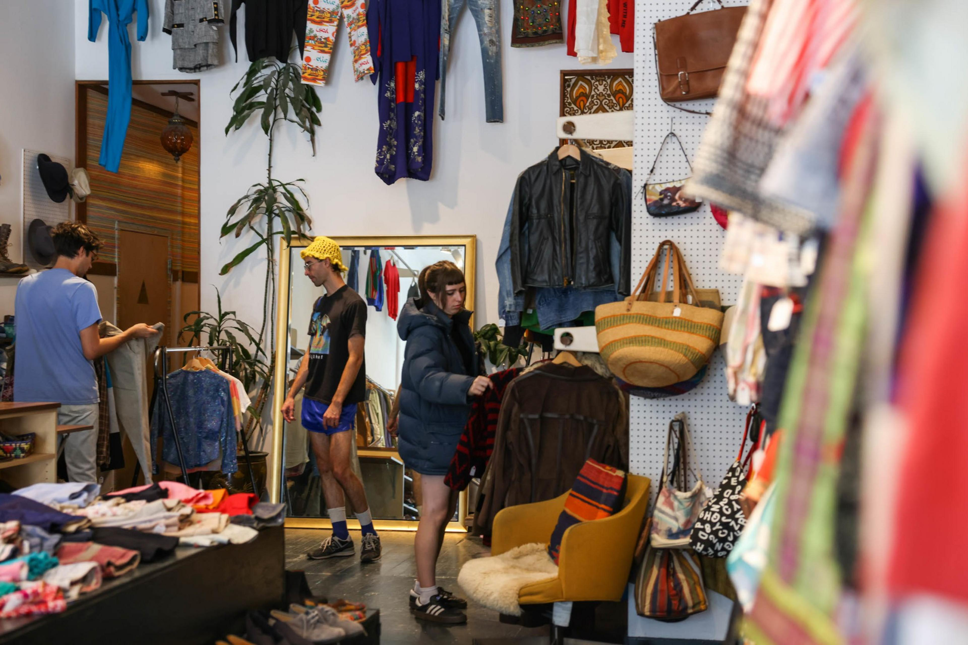 Three people browse in a busy vintage clothing store with jackets, bags, and various clothes displayed on walls, racks, and tables.