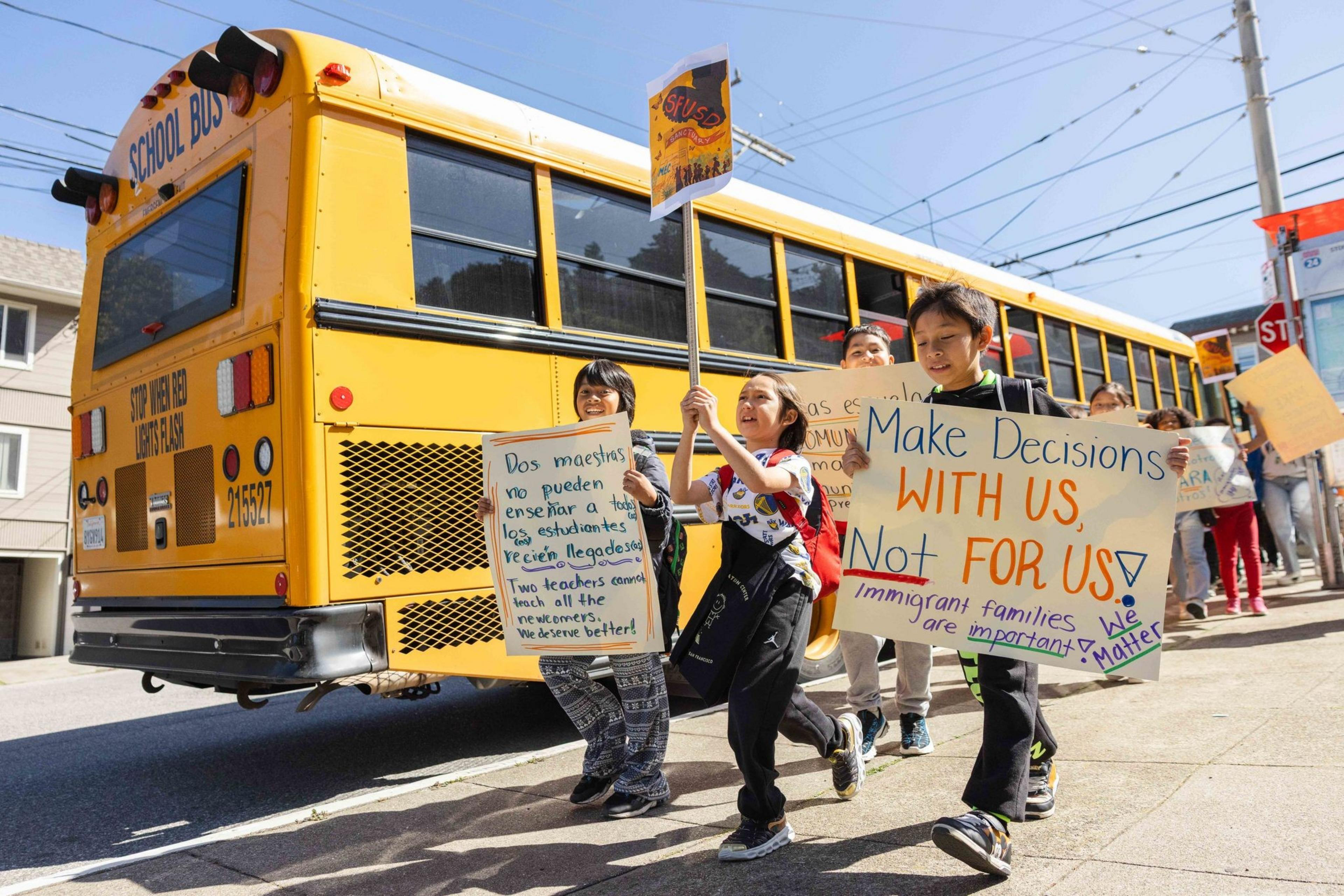 Children hold protest signs about education beside a yellow school bus. The signs express demands for inclusive decision-making and support for immigrant families.
