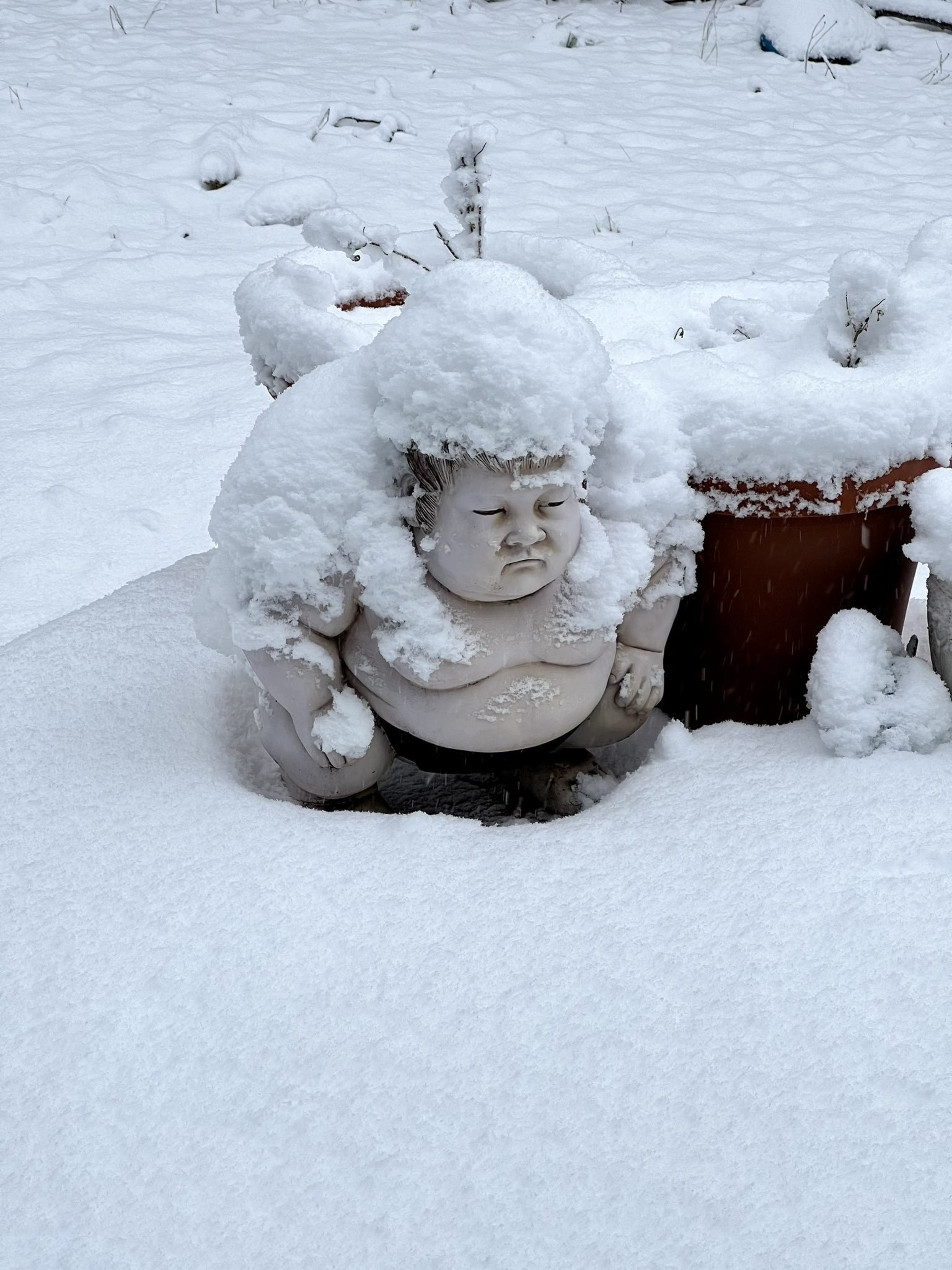 A snow-covered sumo wrestler statue crouches beside a snow-covered plant pot in a snowy garden.
