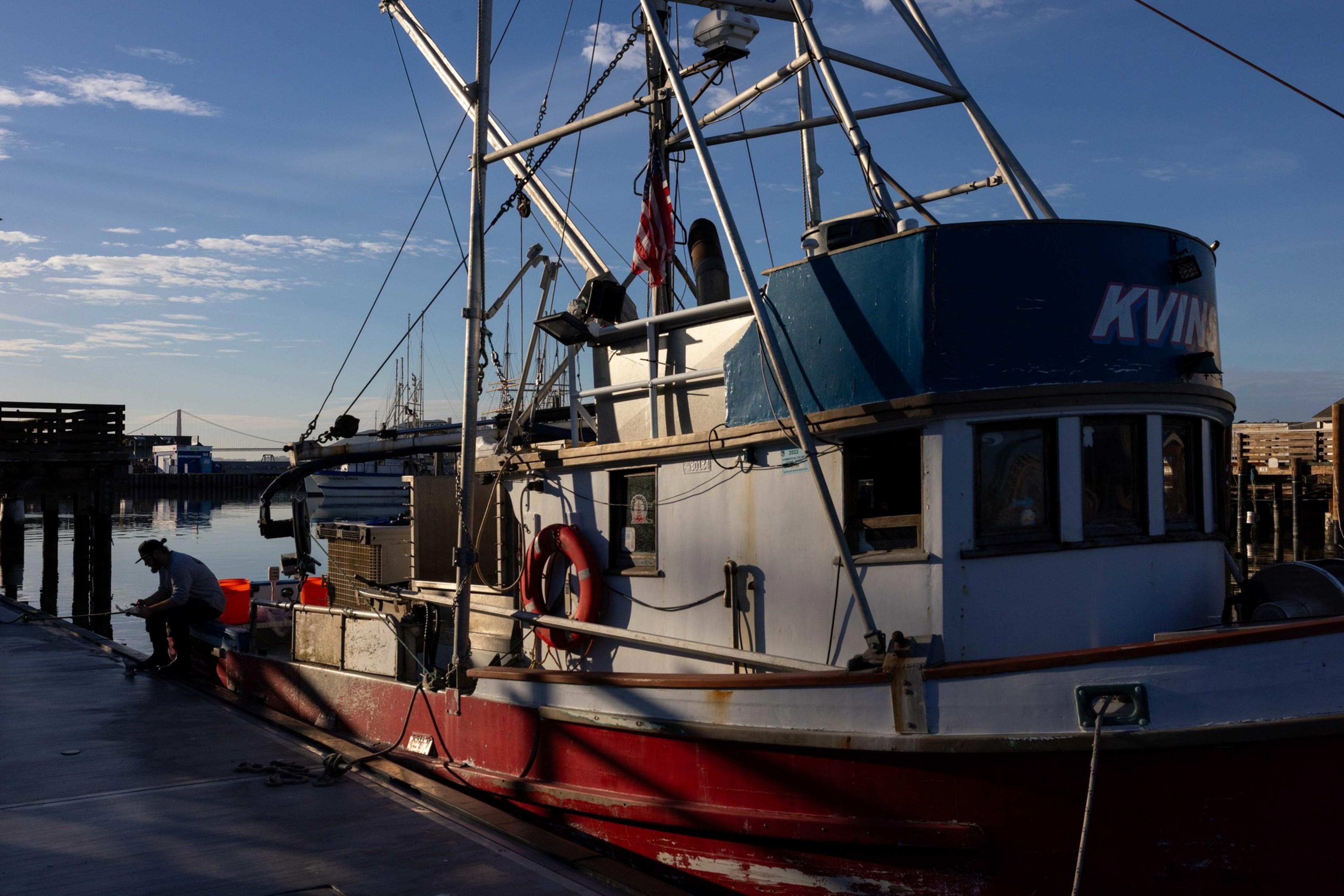A fishing boat is docked at a pier, with a person working beside it. The boat has a blue and red hull, and there's an American flag on the mast.