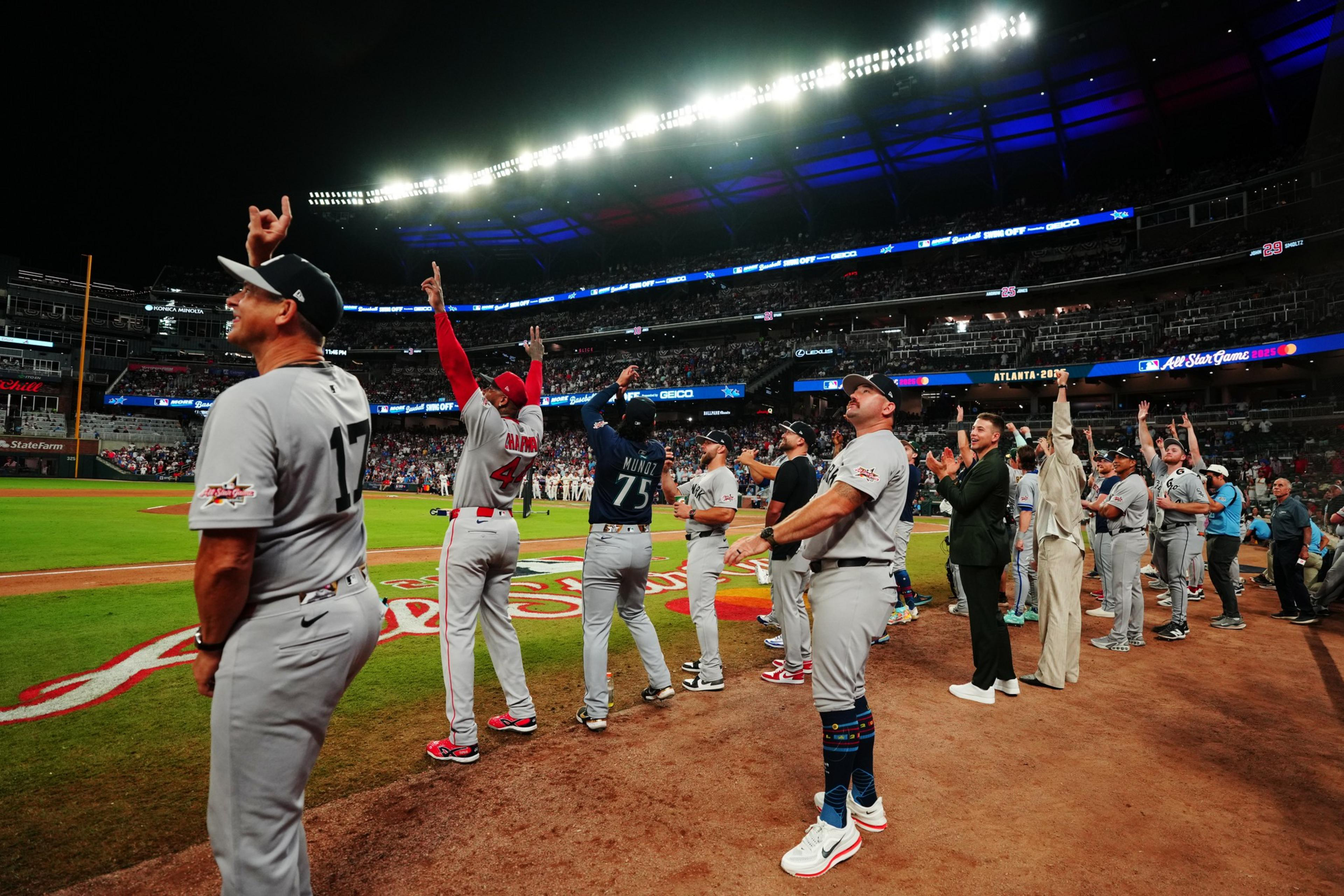 A group of baseball players and staff are on the field, looking and pointing upwards with excitement. The stadium is packed with spectators under bright lights.