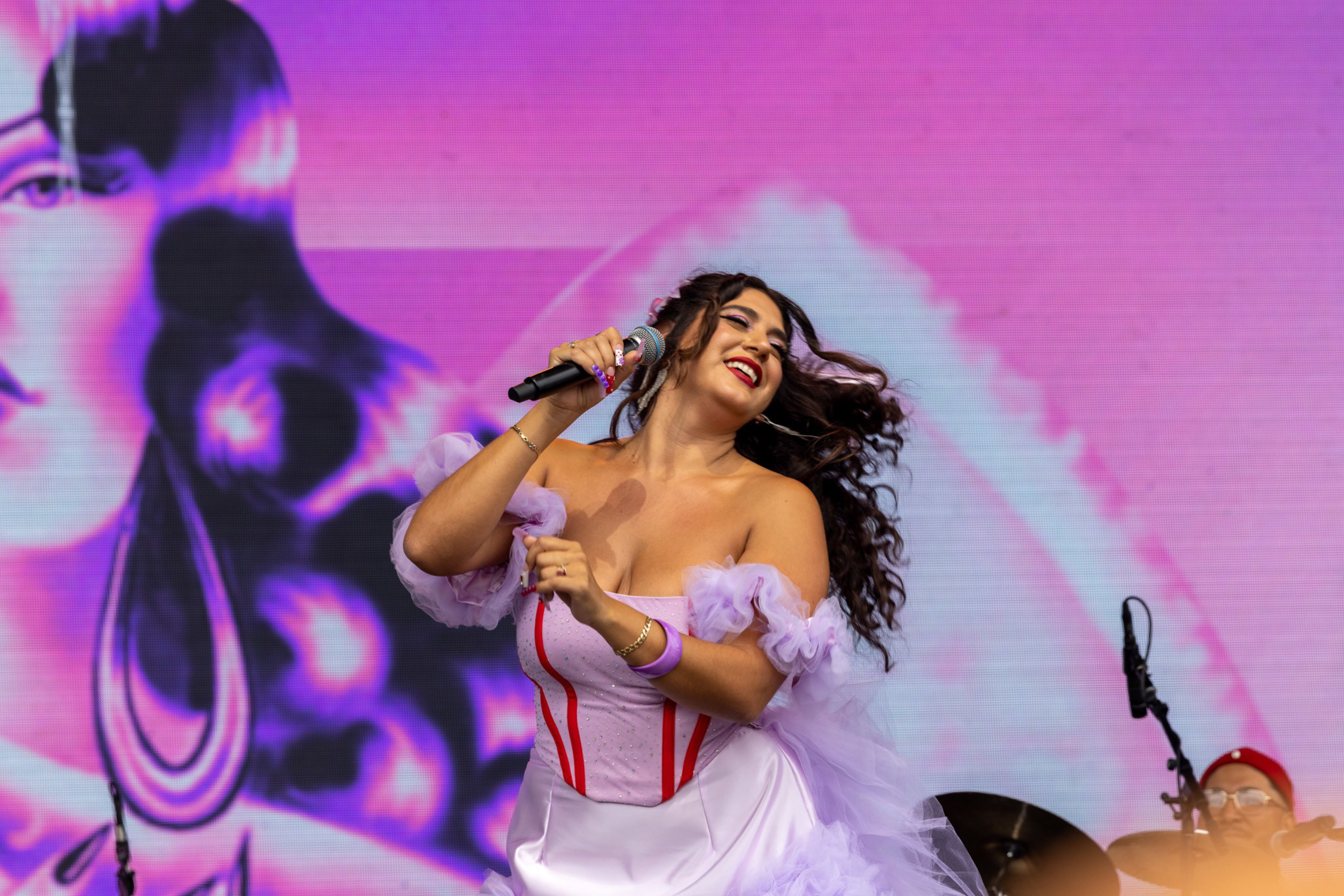 A woman in a flowing lavender dress sings joyfully into a microphone, with her hair blowing and a vibrant purple backdrop behind her.