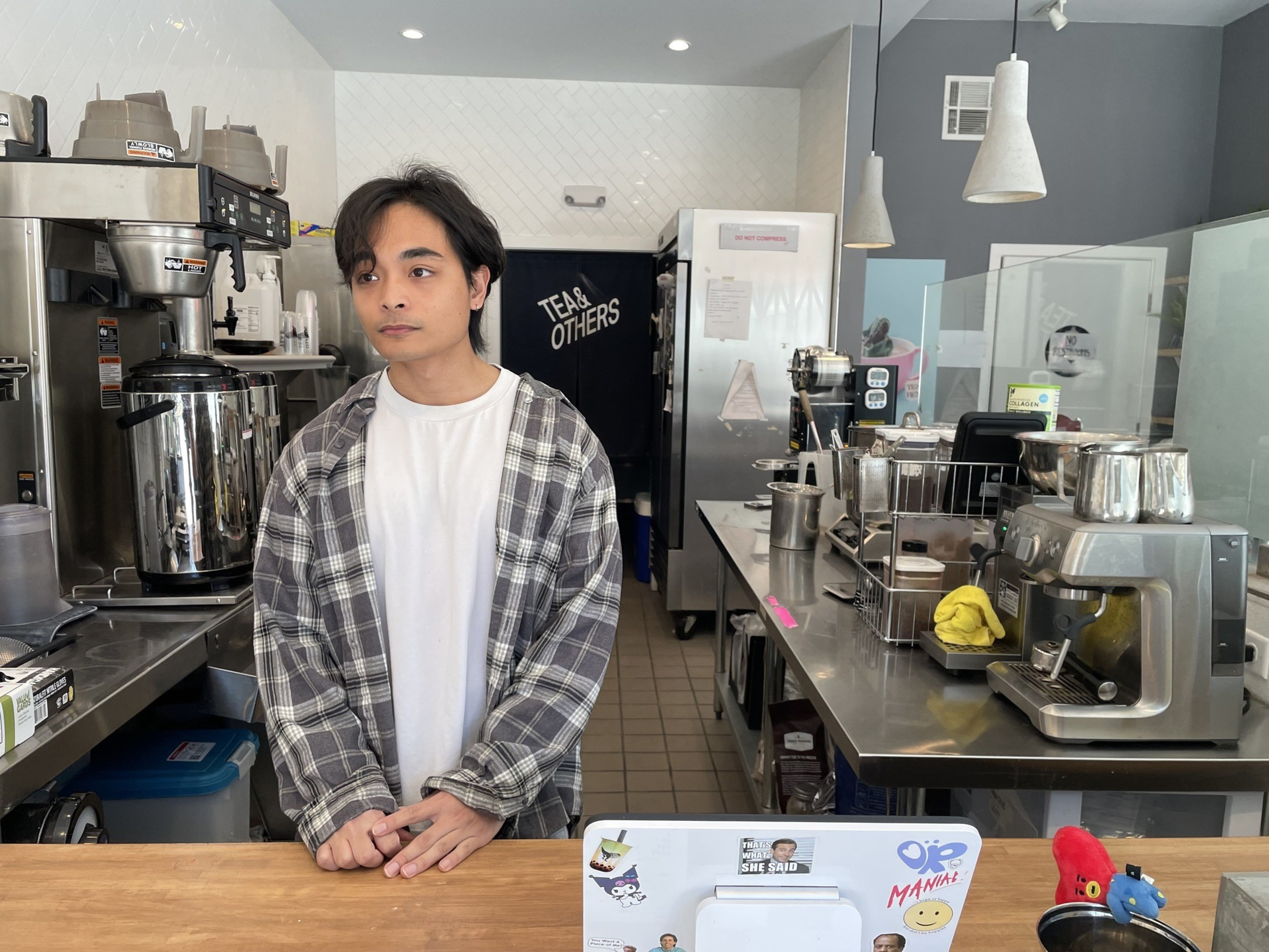 A person stands behind a cafe counter with coffee equipment and lights overhead.