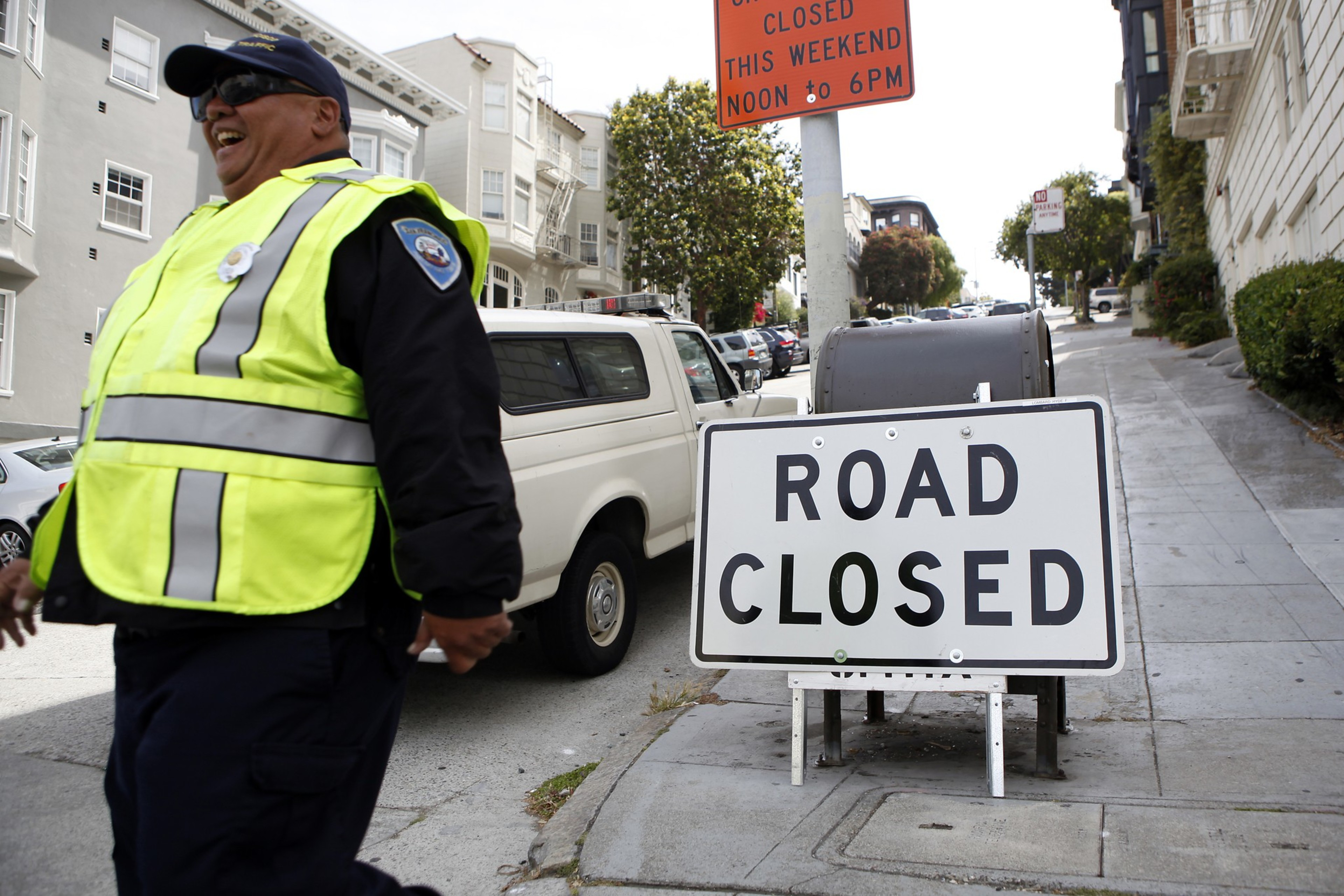 A smiling police officer in a neon yellow safety vest stands near a "ROAD CLOSED" sign on a city sidewalk with parked cars and buildings in the background.
