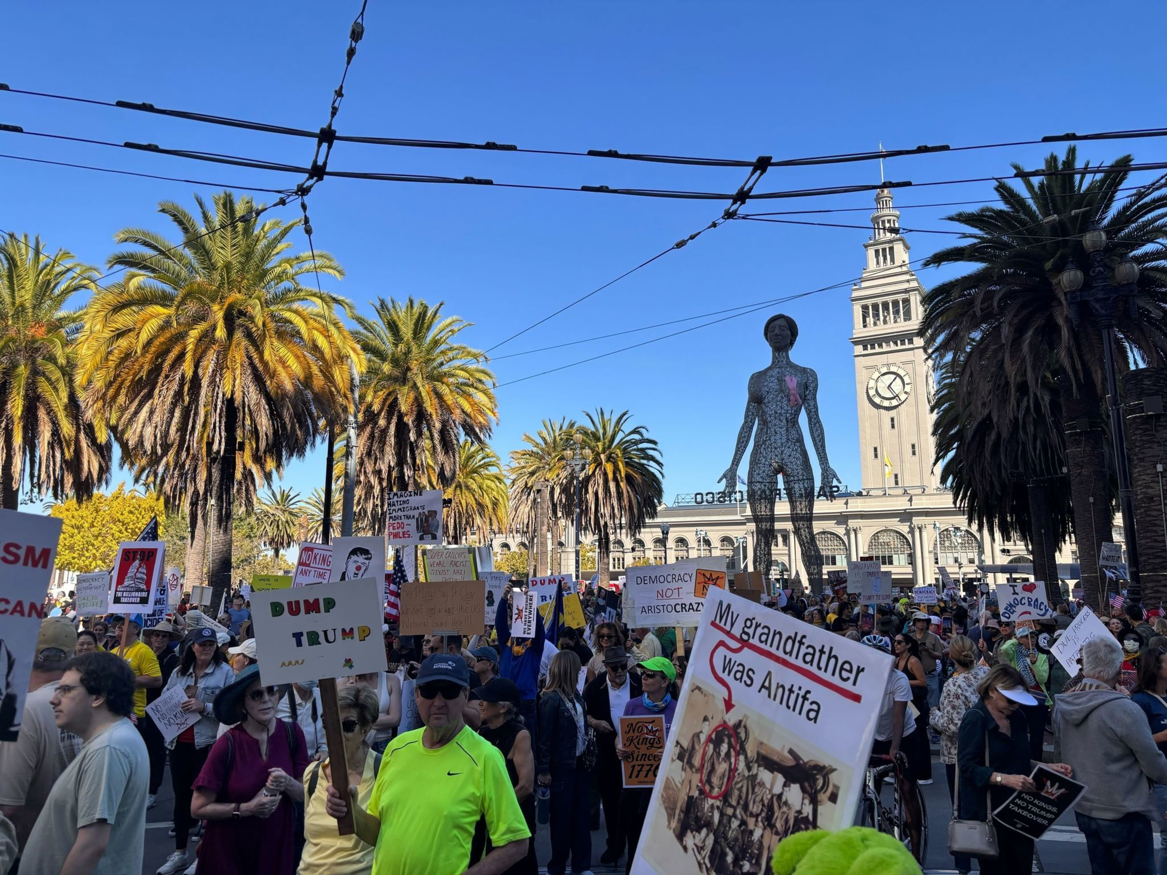 A large crowd holds protest signs including "Dump Trump" and "My grandfather was Antifa" near palm trees and a tall clock tower under a clear blue sky.