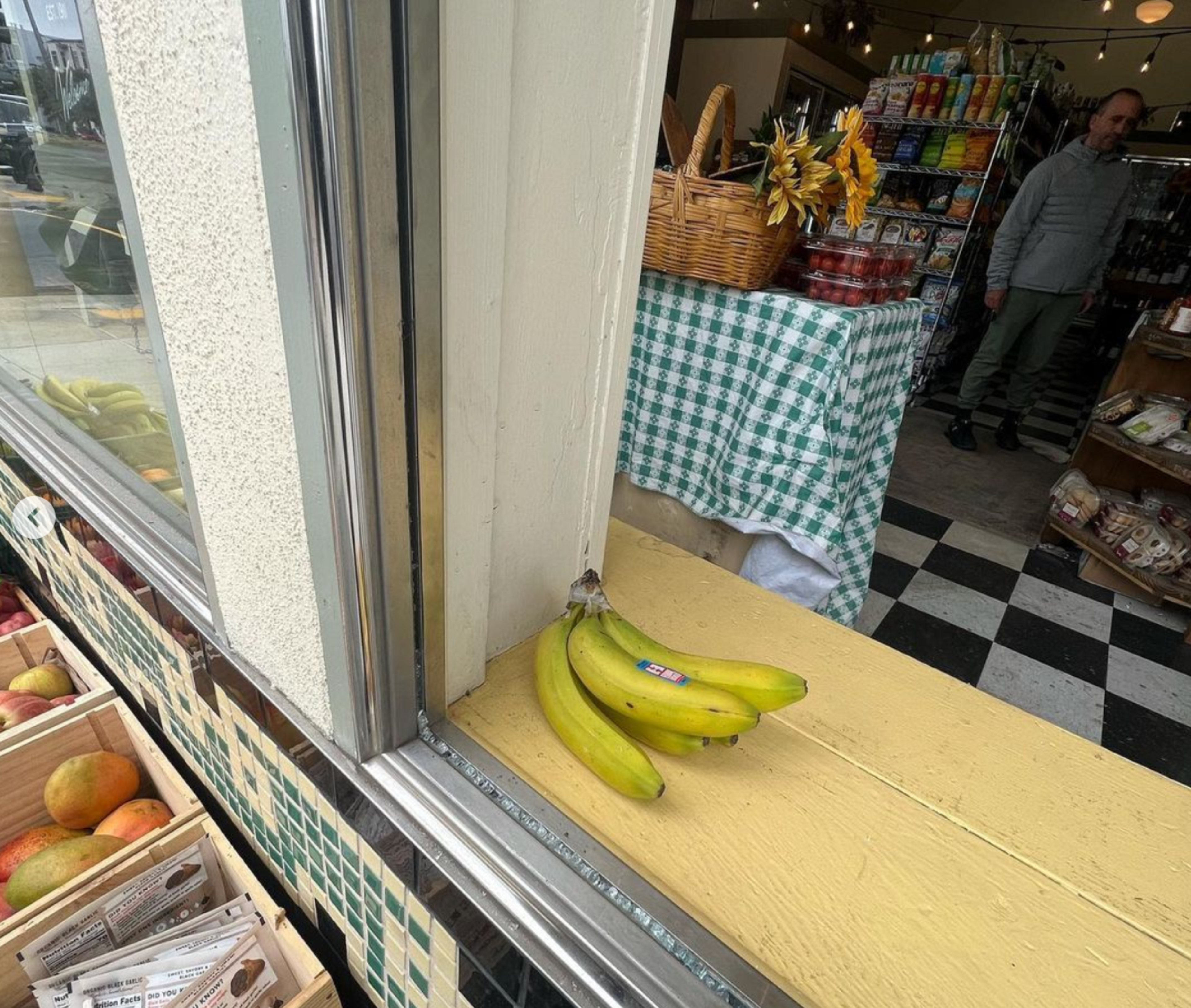A bunch of bananas rests on a yellow windowsill with a checkered green-and-white tablecloth and a basket with sunflowers inside a store.