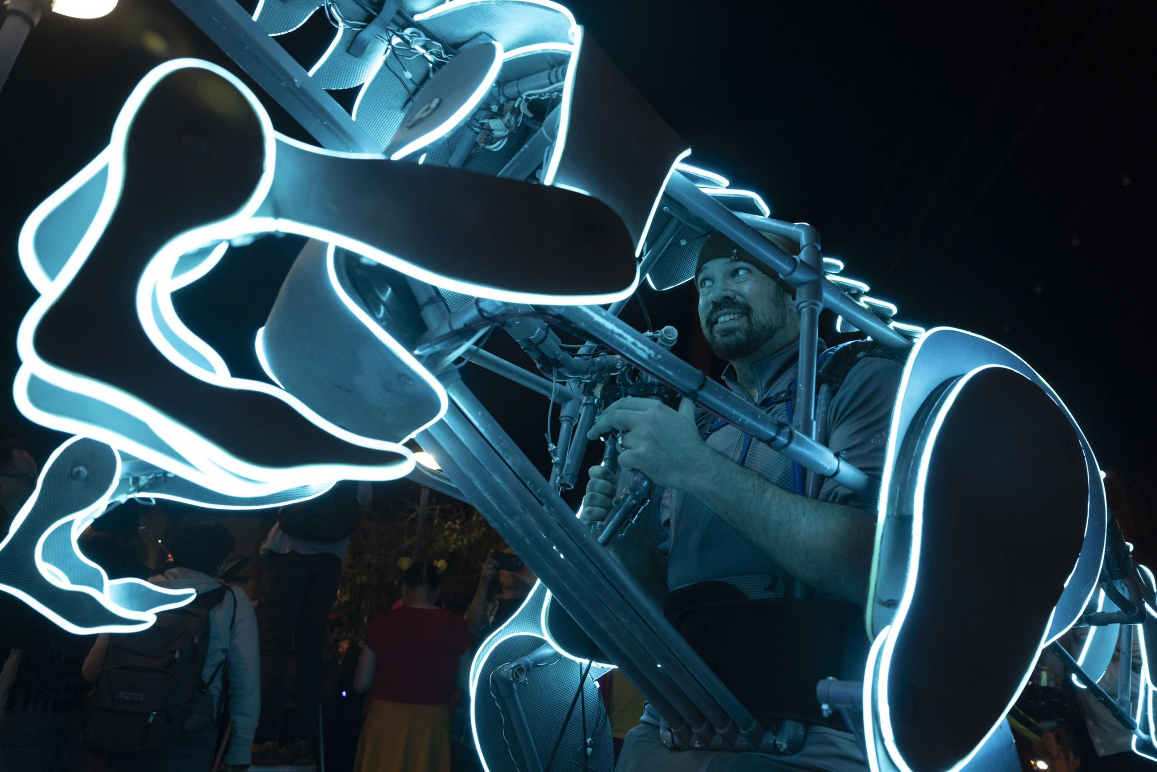 A person smiles while playing an instrument within a glowing, neon-lit structure at night. The abstract design creates a vibrant, futuristic feel.