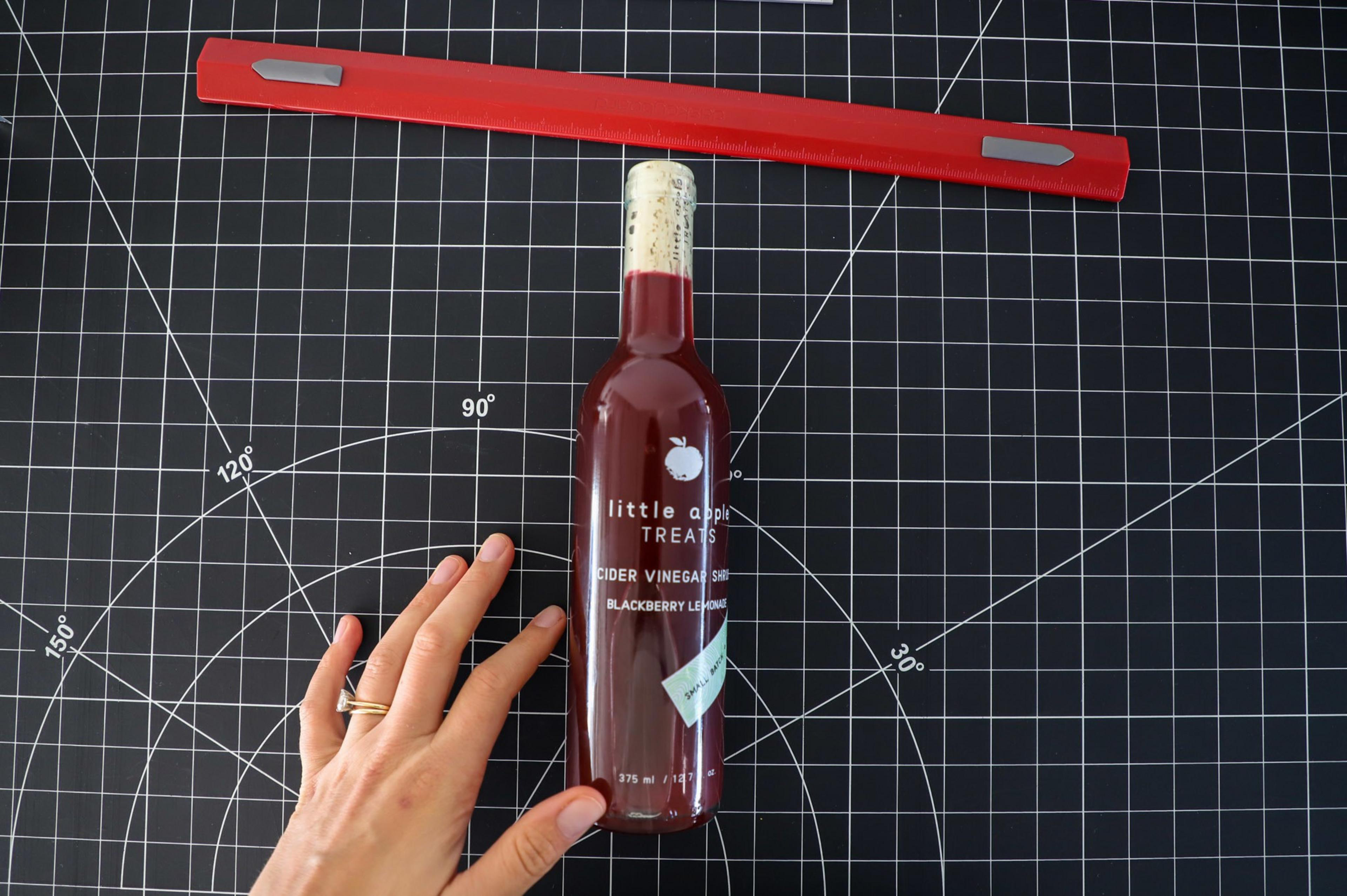 A bottle of purple blackberry shrub with a cork at the top rests on a table, with the author's hand next to it for scale.