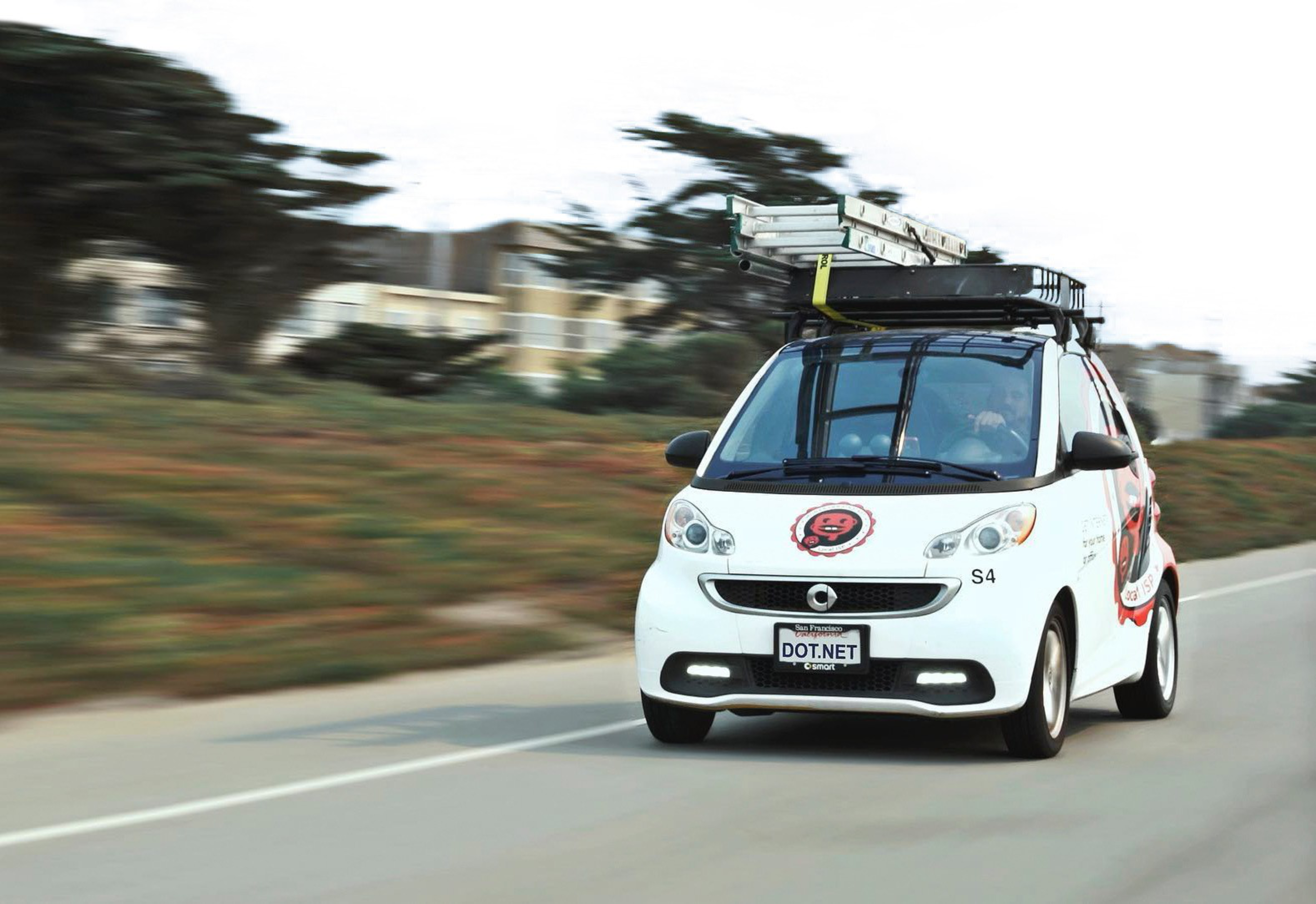 A small white car with roof equipment and decals drives along a road with greenery.