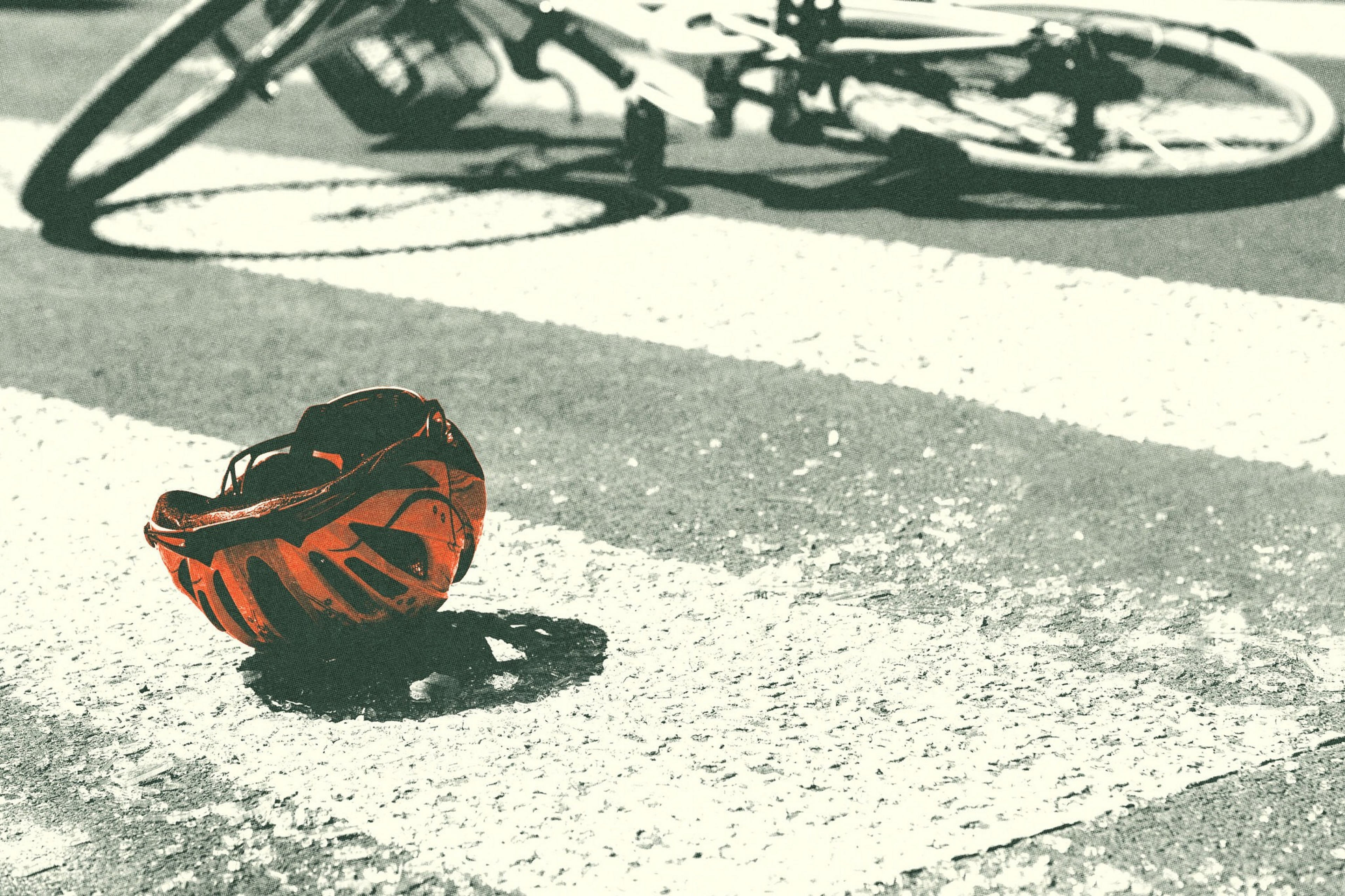 A red bicycle helmet lies on a crosswalk, with a fallen bicycle blurred in the background, suggesting a recent accident.