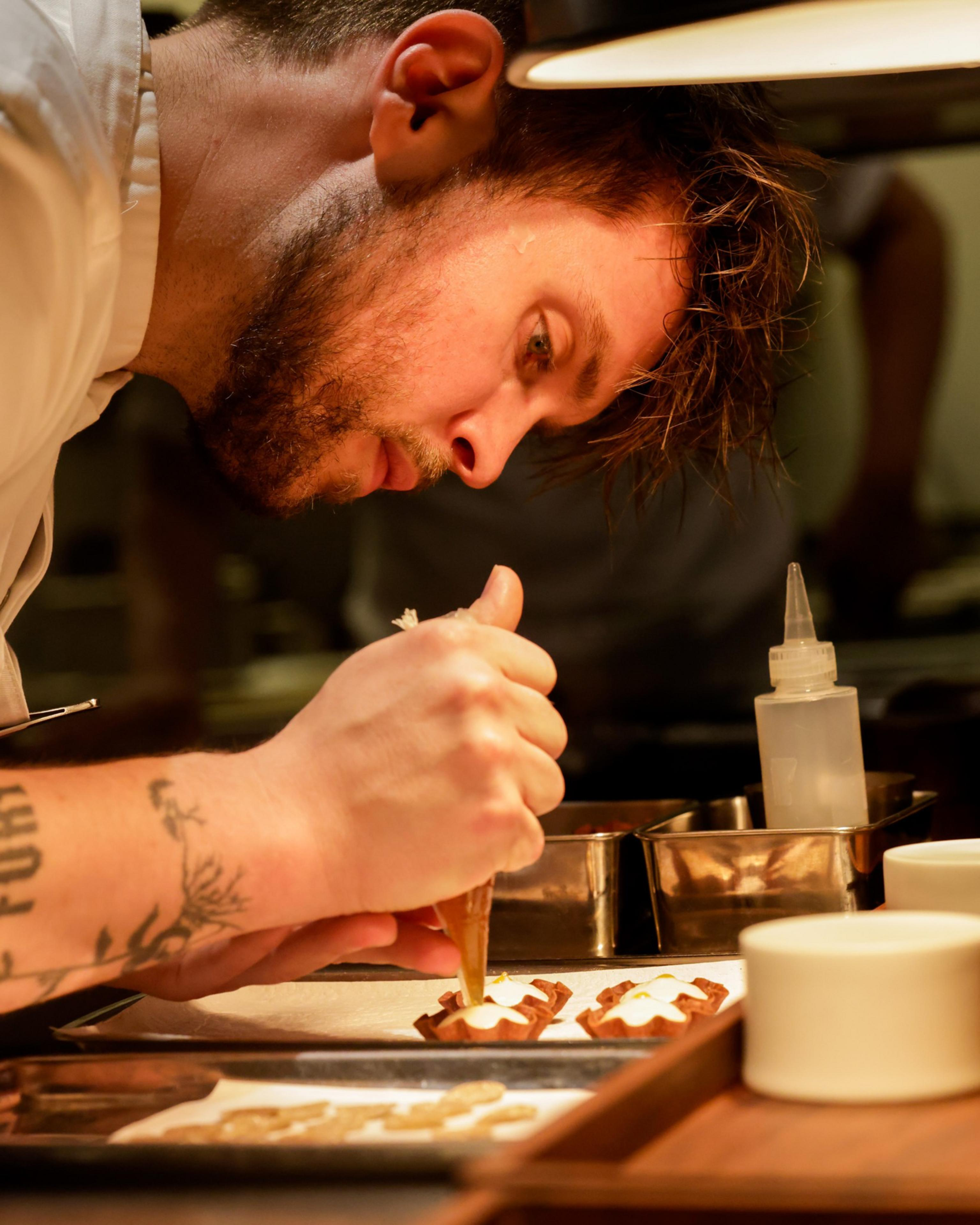 A chef with a tattooed arm is focused on piping cream onto small pastry shells in a kitchen, with various containers and cooking tools around.