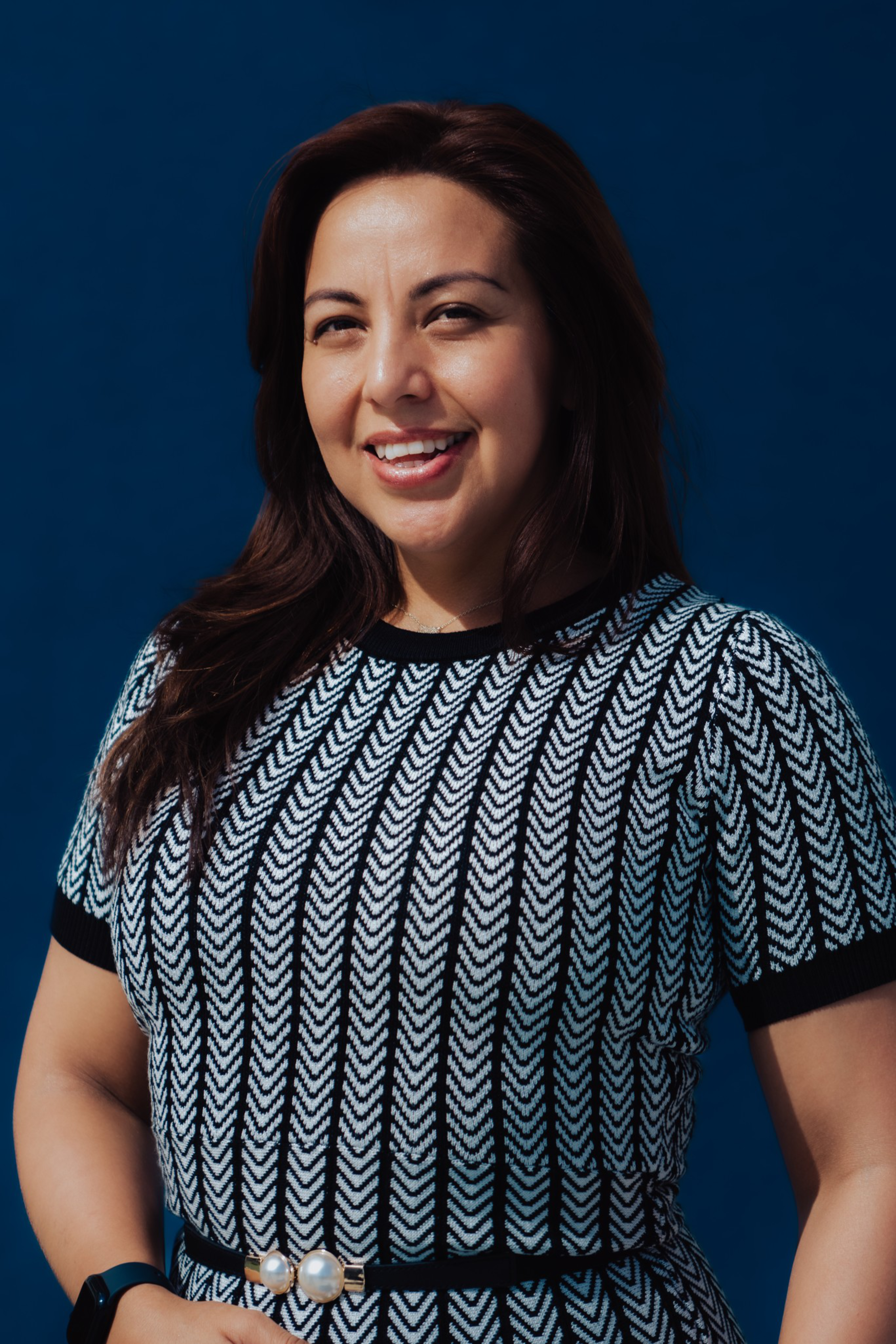 A smiling woman with long dark hair wears a black and white patterned dress with a pearl-accented belt, standing against a deep blue background.