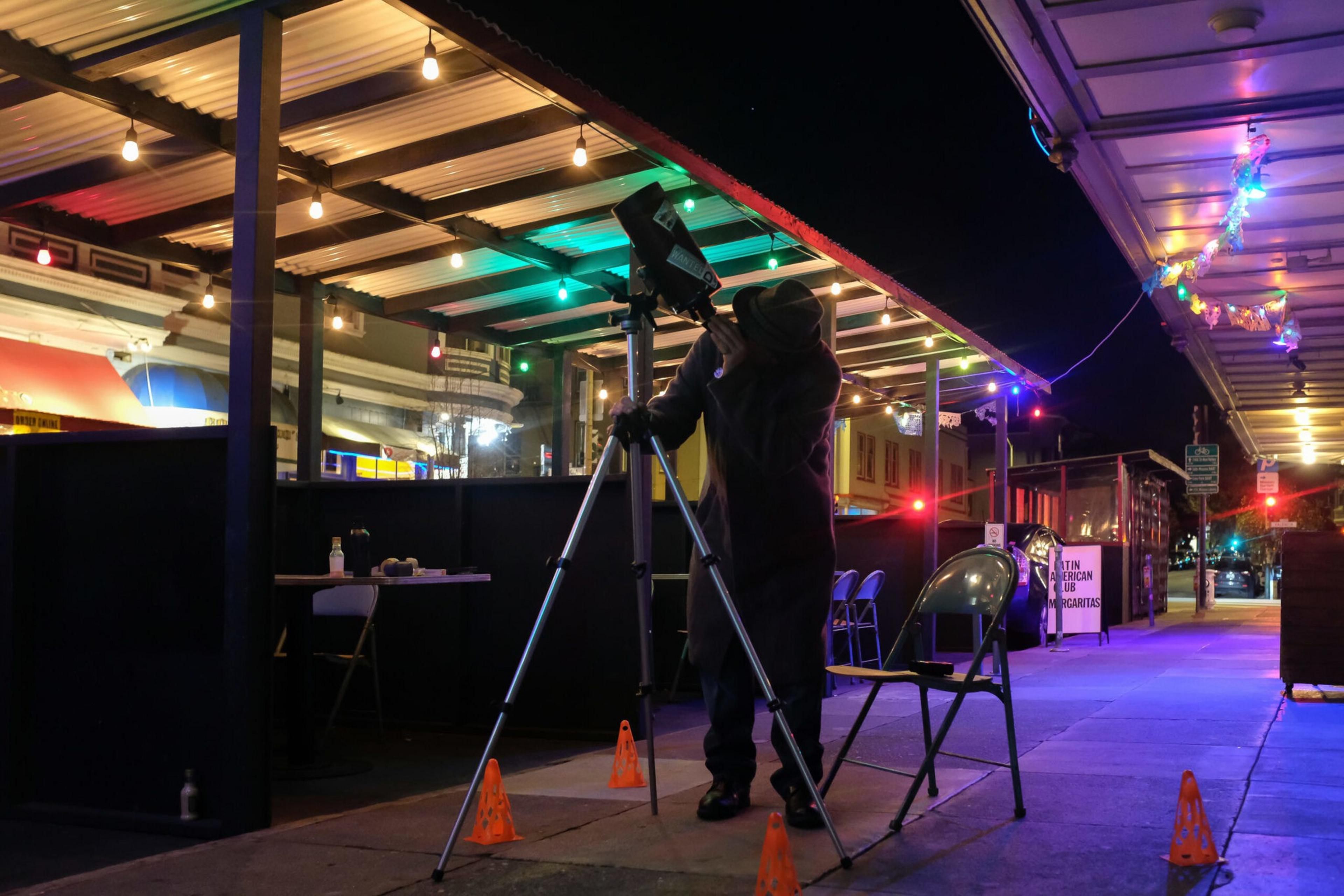A person in a coat and hat adjusts a camera on a tripod under outdoor string lights on a nighttime sidewalk with orange cones around.