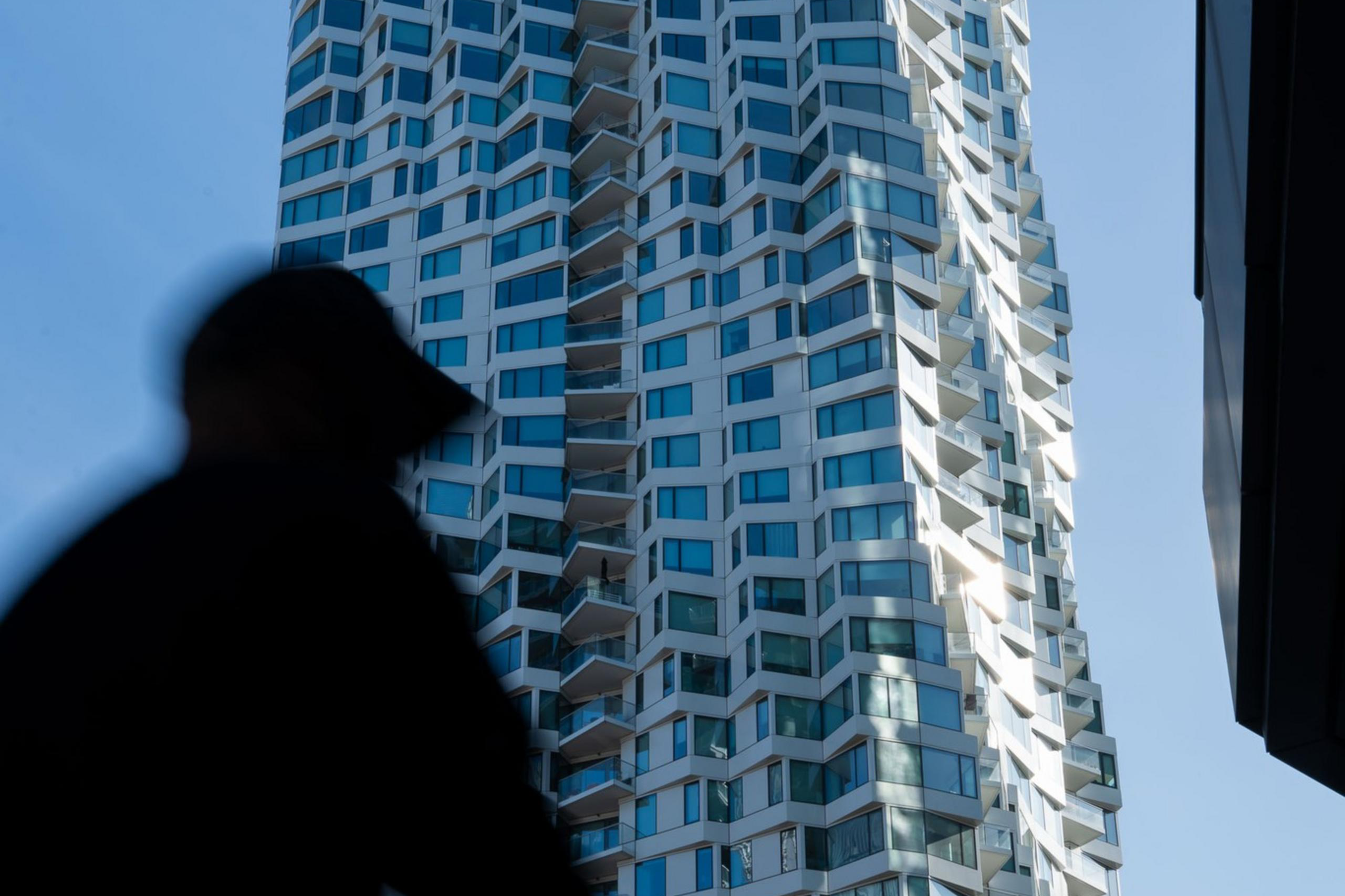 A tall, modern skyscraper with a wavy, geometric design stands against a blue sky. In the foreground, a blurred silhouette of a person is visible.