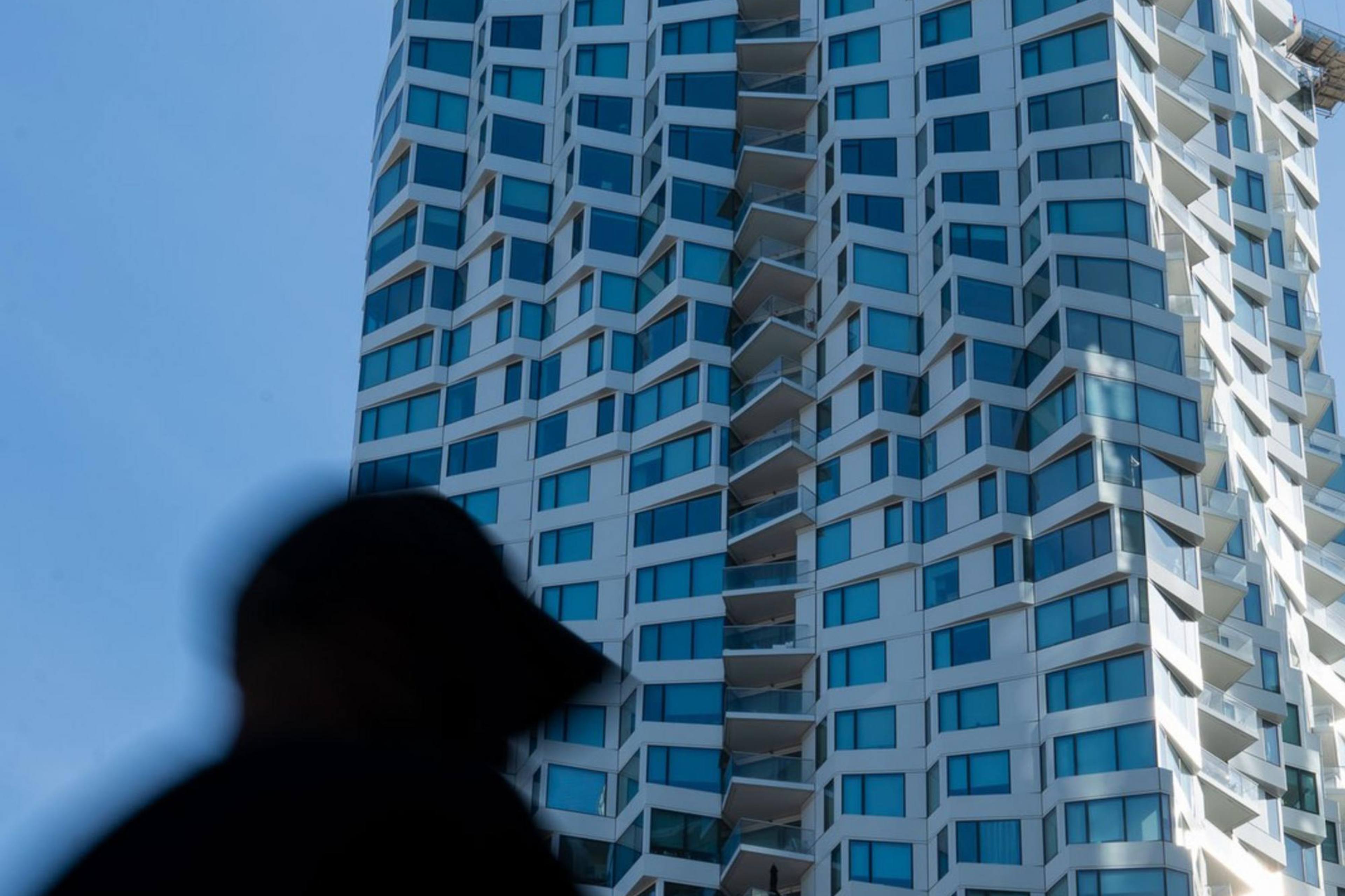 A tall, modern skyscraper with a wavy, geometric design stands against a blue sky. In the foreground, a blurred silhouette of a person is visible.
