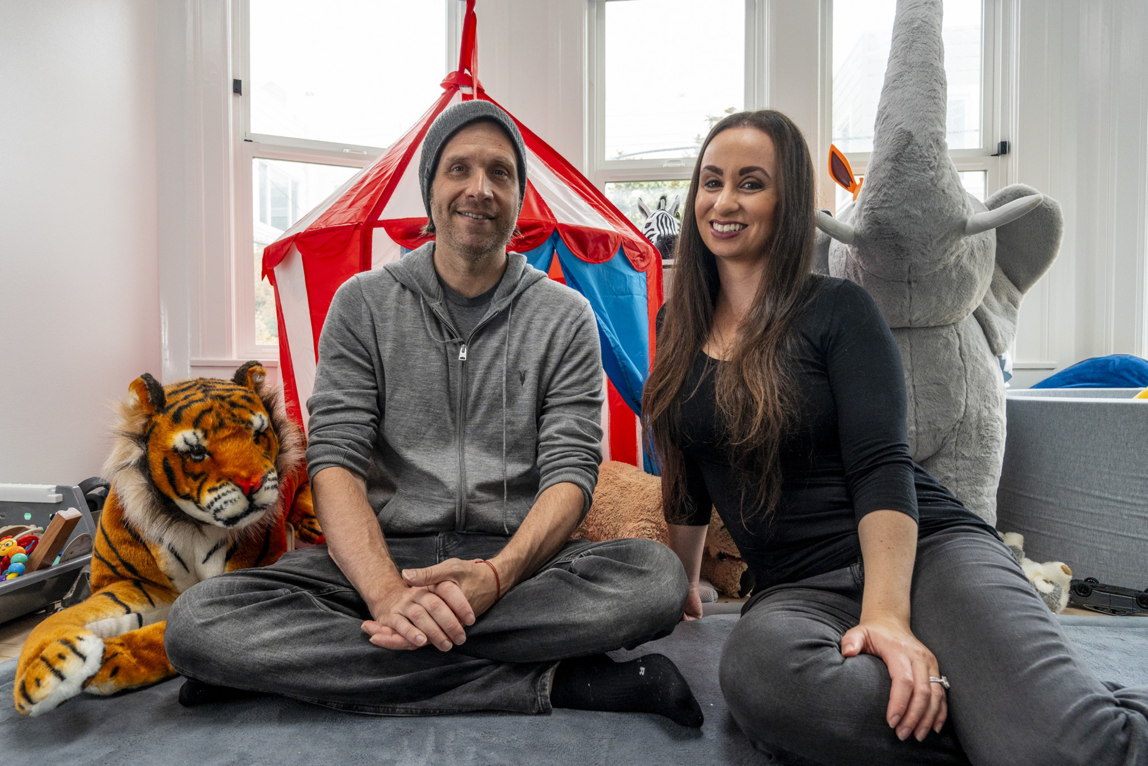 A man and woman sit cross-legged on a carpeted floor between large stuffed tiger and elephant toys, with a red play tent behind them.