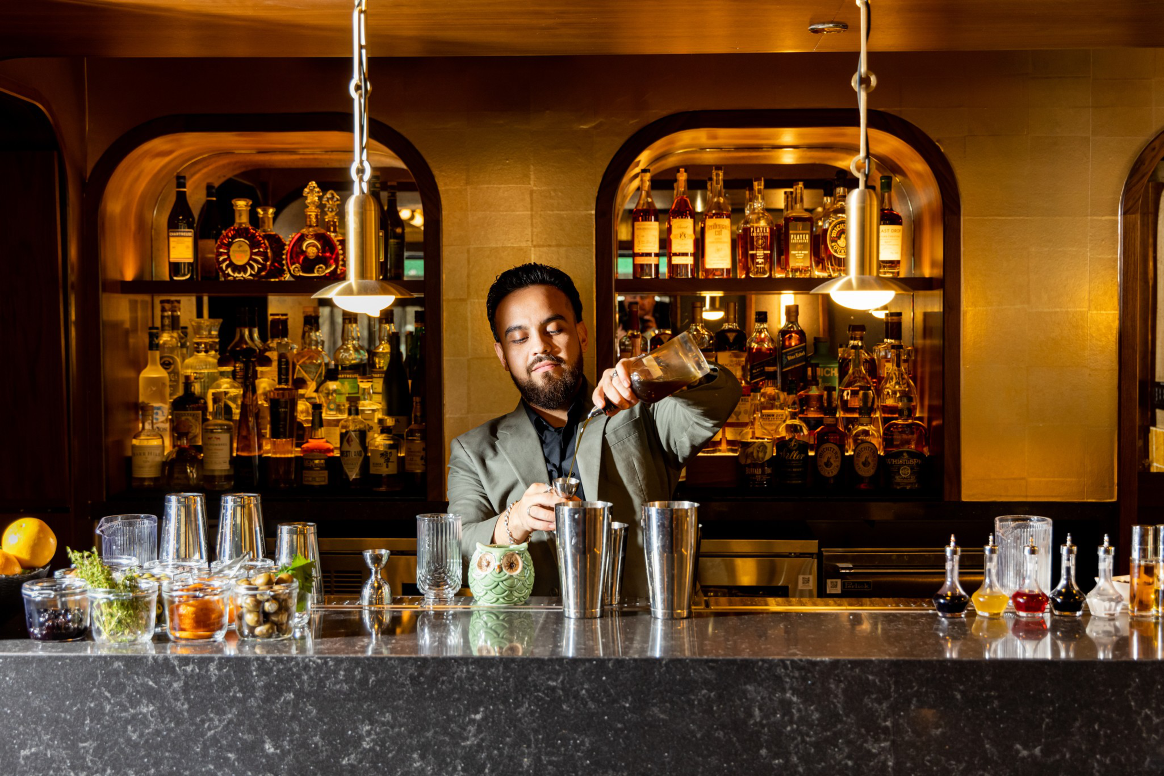 A bartender in a suit pours a drink into a shaker behind a bar with various bottles, jars of ingredients, and cocktail tools on the counter.