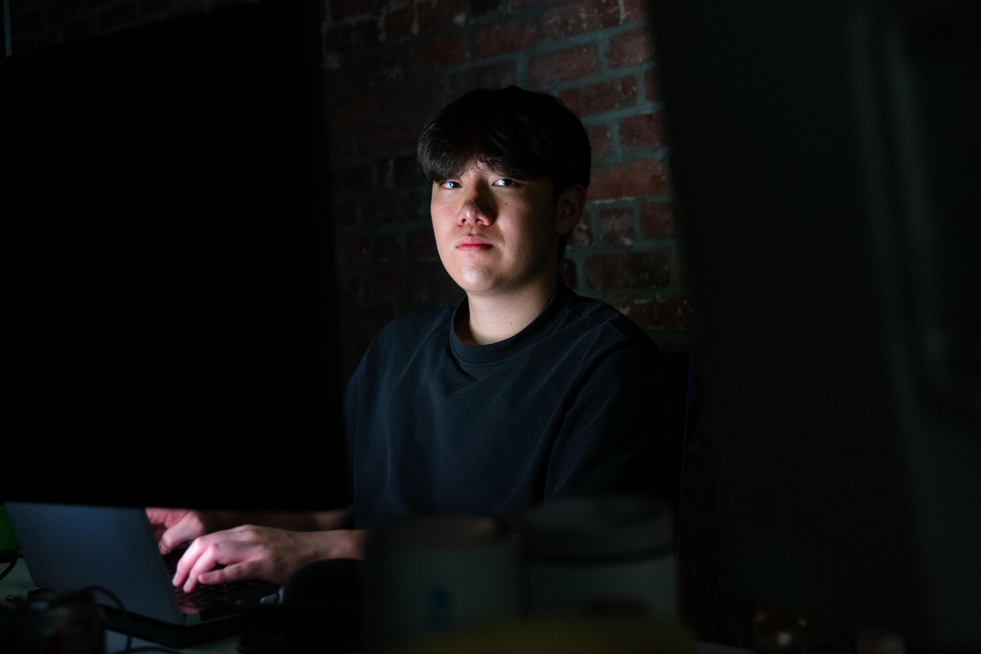 A young man with black hair sits by a laptop in a dimly lit room with a brick wall, looking directly at the camera with a neutral expression.