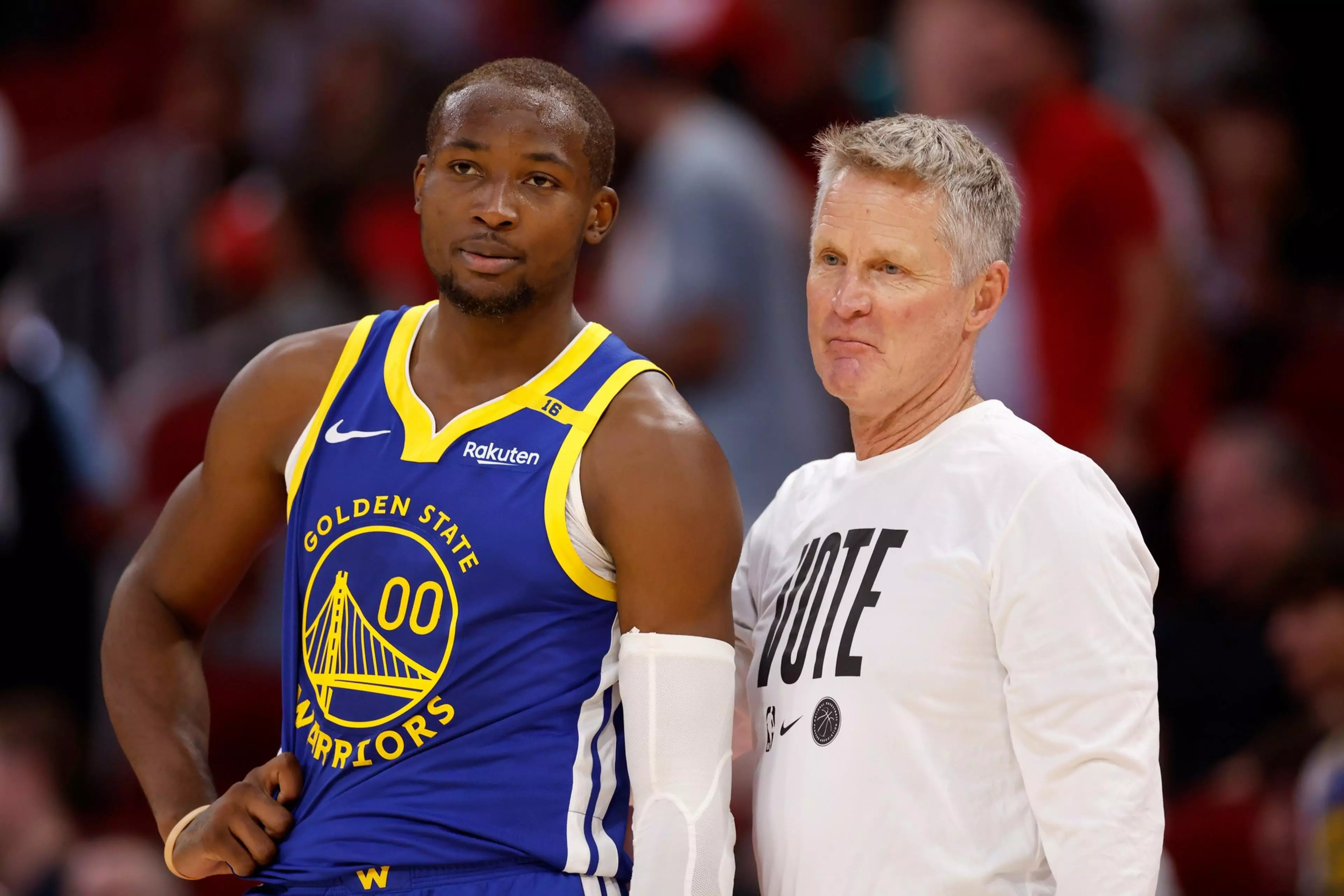 A basketball player in a blue Golden State Warriors jersey stands beside a person in a white shirt with "VOTE" written on it.