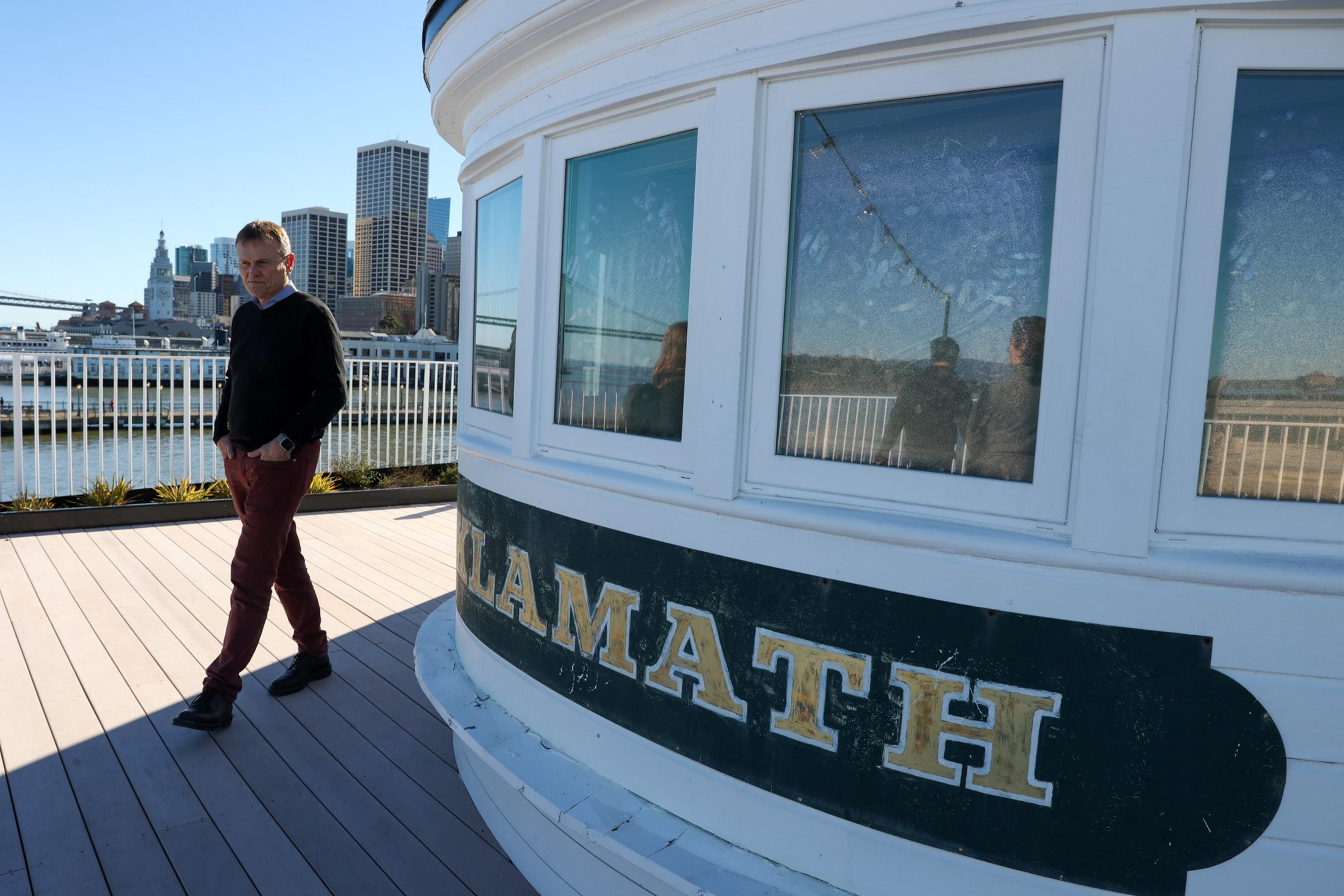 A man in a black sweater and maroon pants walks on a wooden deck near a white boat with "KLAMATH" written on its side, with city buildings in the background.