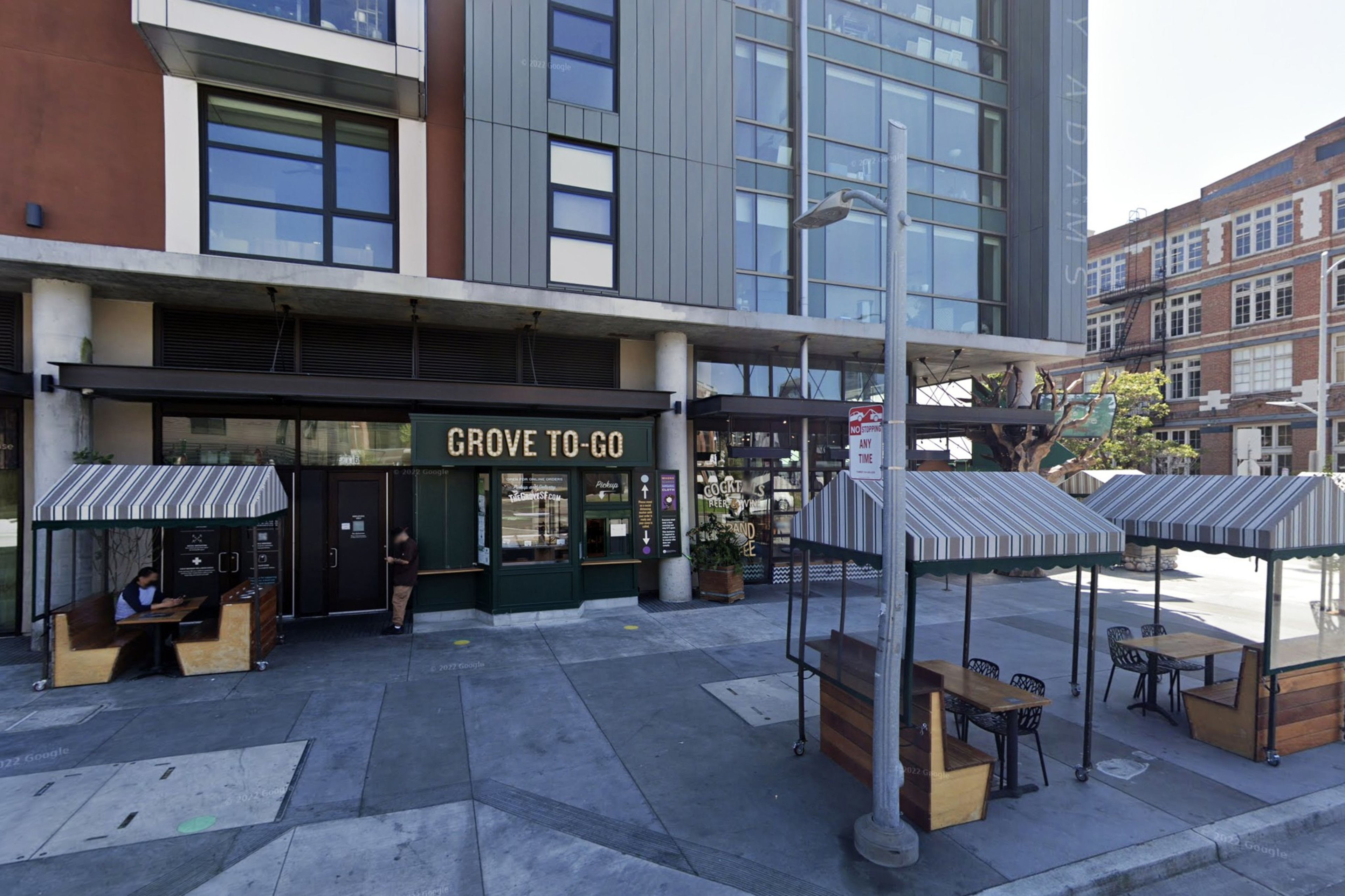 A café named "Grove To-Go" with outdoor seating areas featuring small tables, striped canopies, and wooden benches on a city sidewalk.
