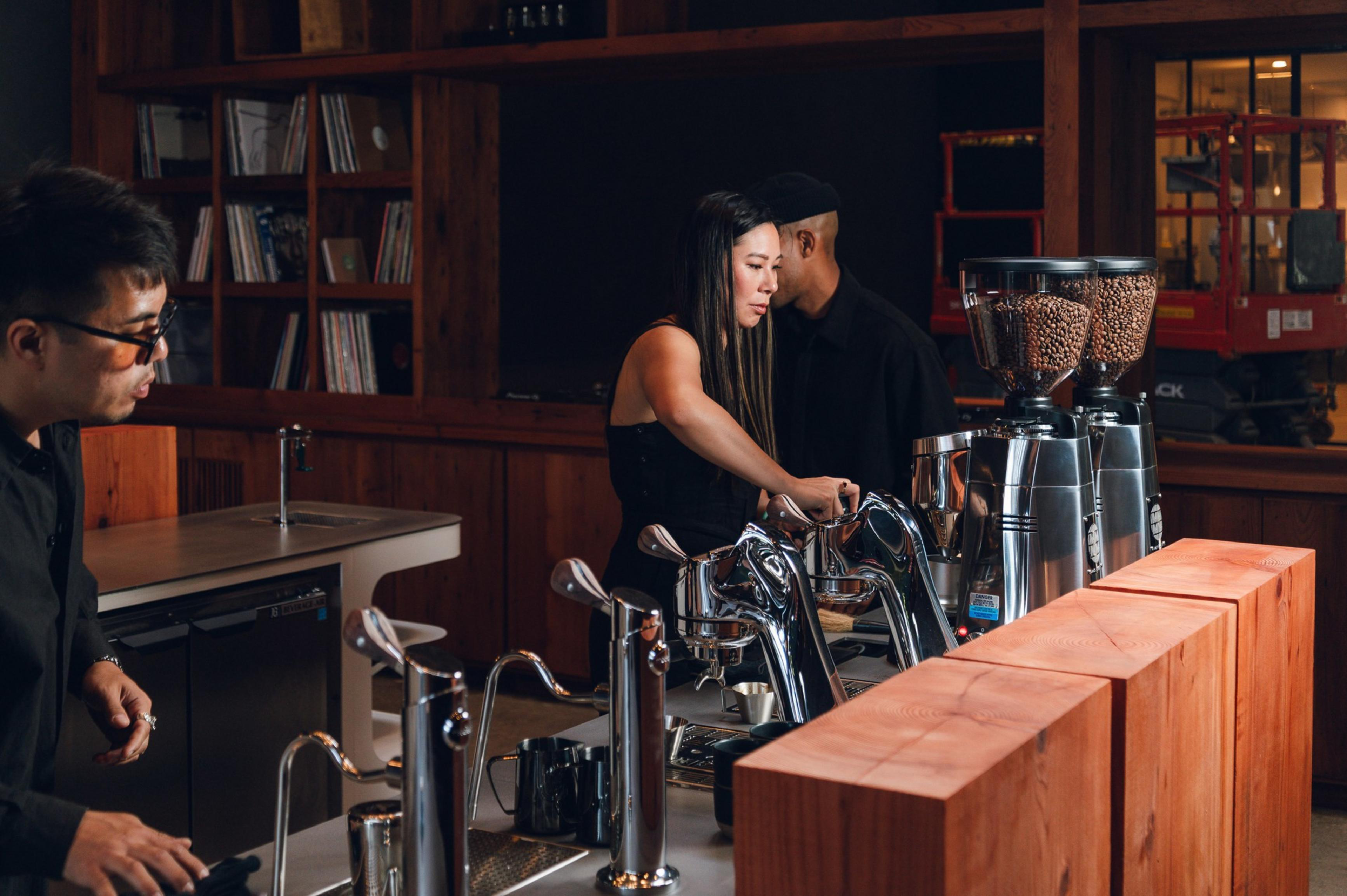 Three people work behind a coffee bar with espresso machines and coffee grinders, set against wooden shelves and warm, dim lighting.