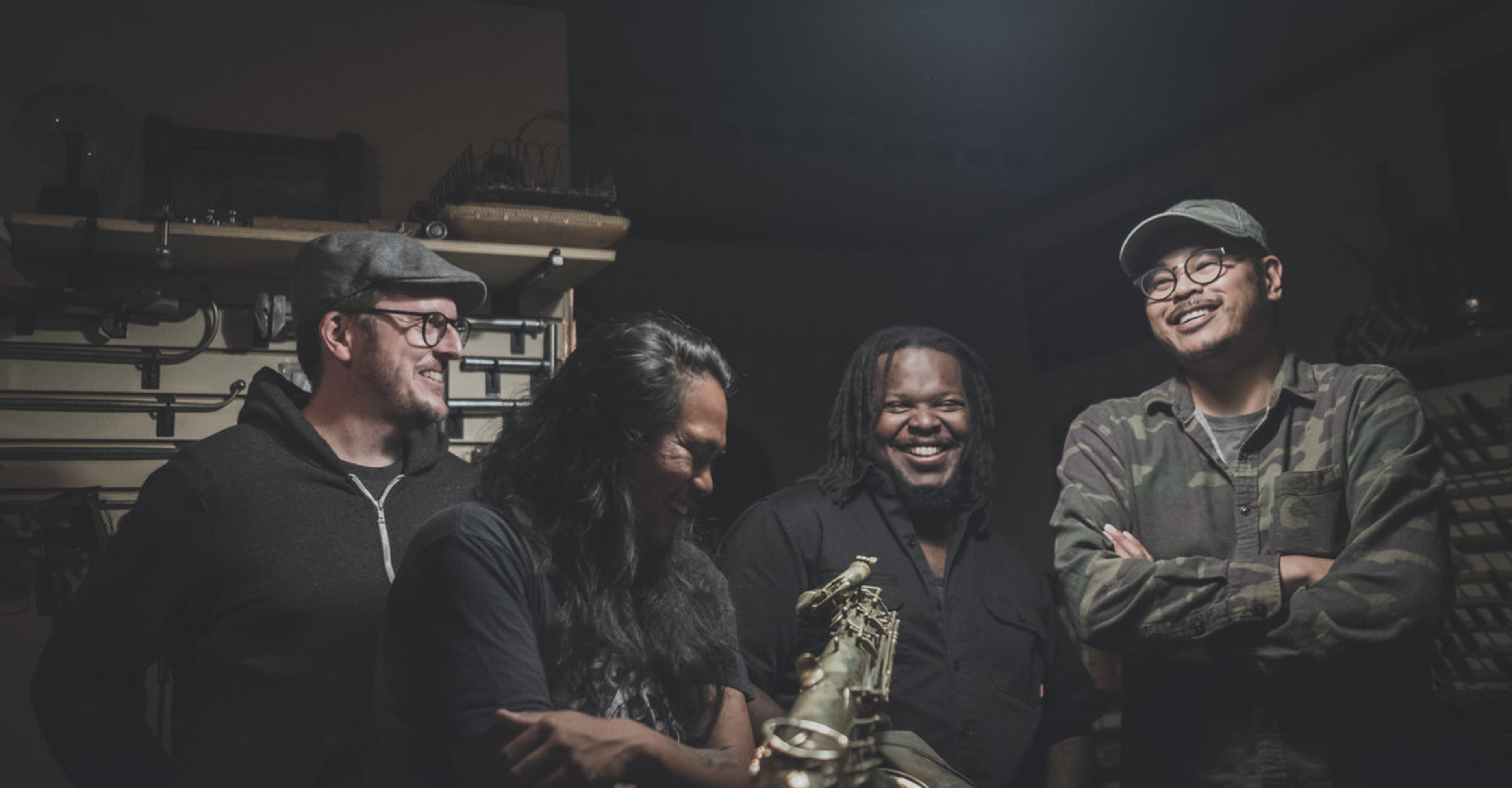 Four men stand closely together in a dimly lit room, smiling and laughing, with one holding a saxophone and various tools visible in the background.