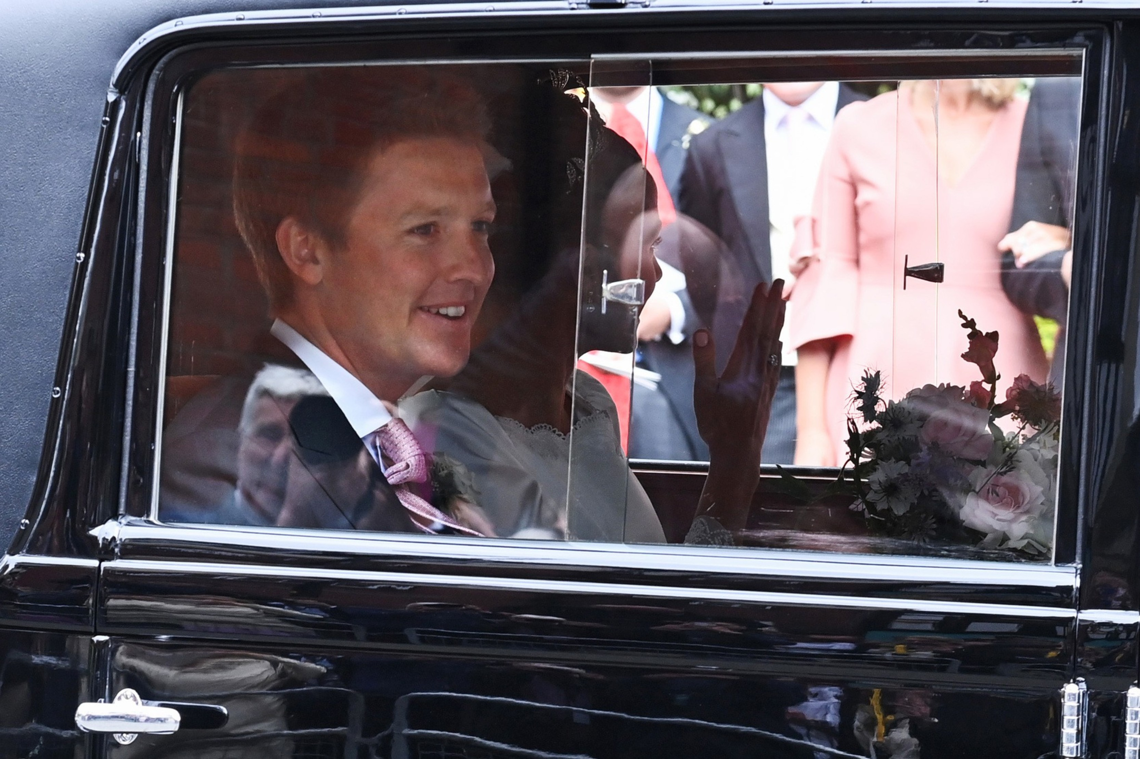 A smiling man in a suit and tie sits next to a woman waving inside a black vintage car with a bouquet of flowers on the seat.