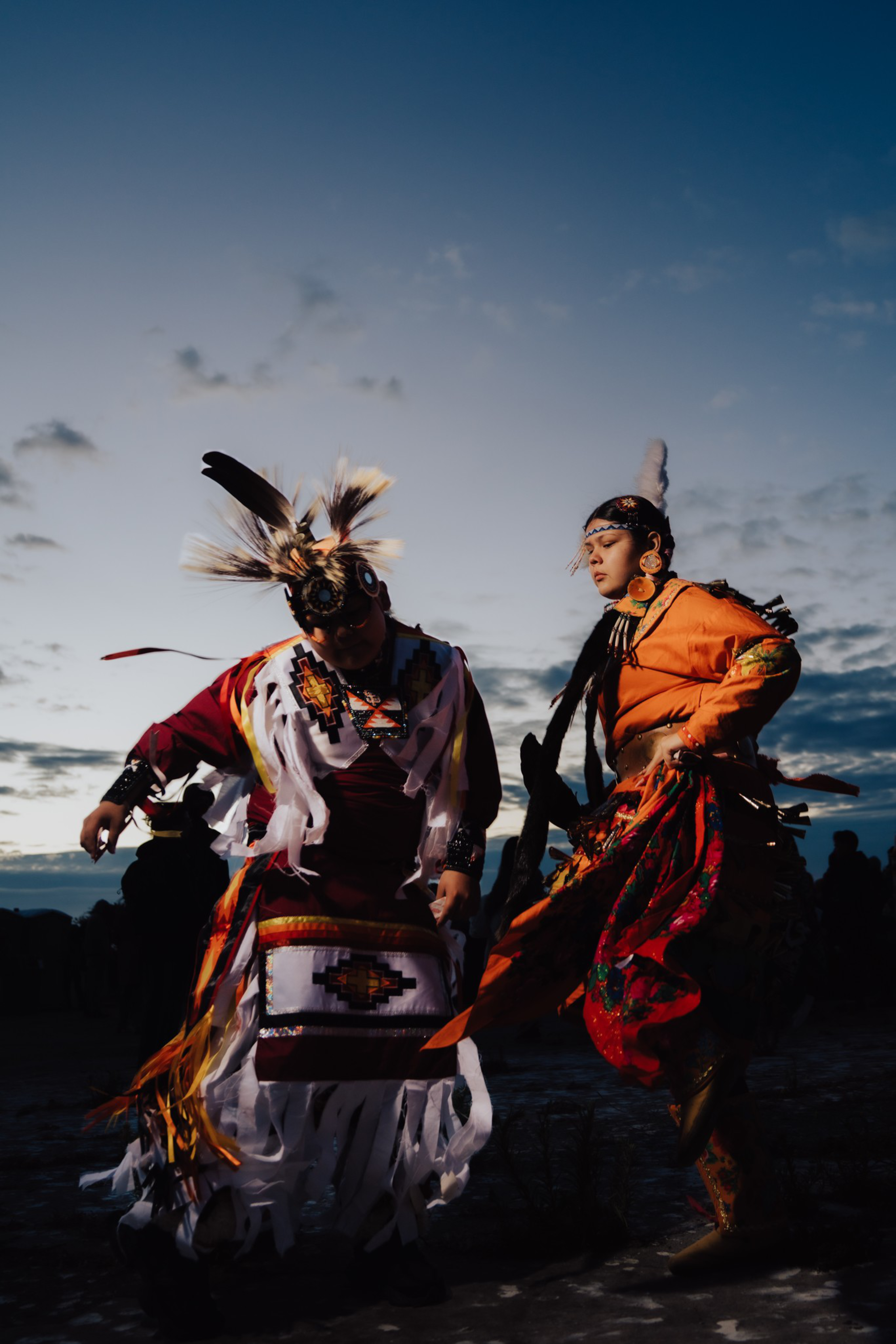 Two people wearing traditional Native American regalia, including feathers and intricate beadwork, dance outdoors at dusk against a cloudy sky.