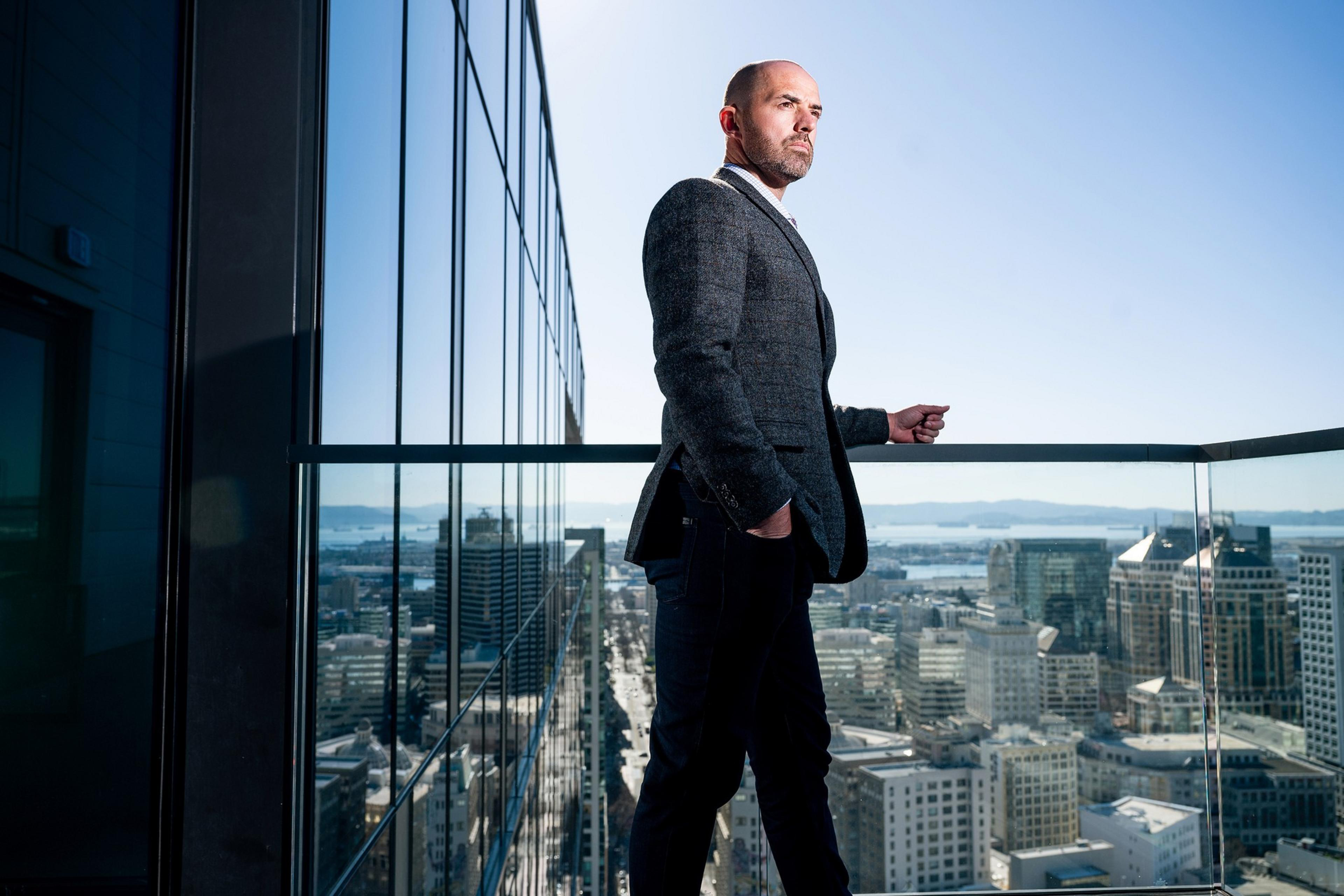 A man in a suit stands confidently on a glass balcony overlooking a cityscape with distant hills and water under a clear blue sky.
