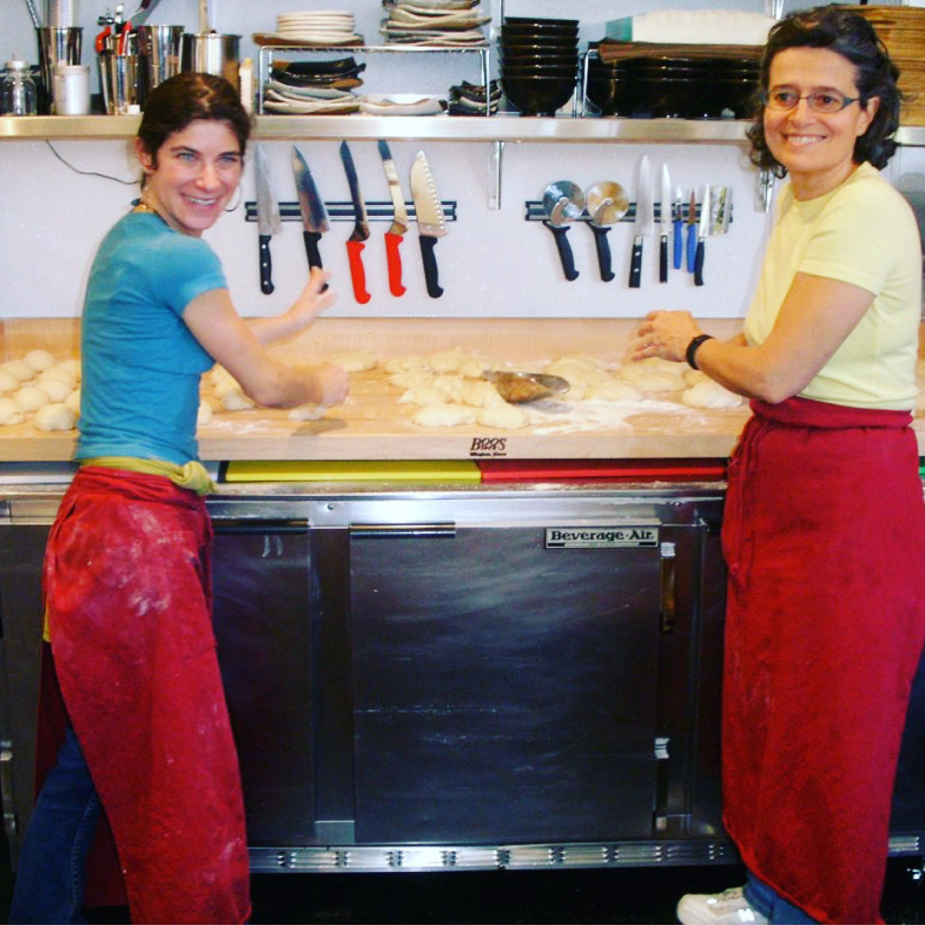 Two women stand at a kitchen counter making dough. They wear colorful shirts and red aprons. Knives and utensils hang on the wall behind them.